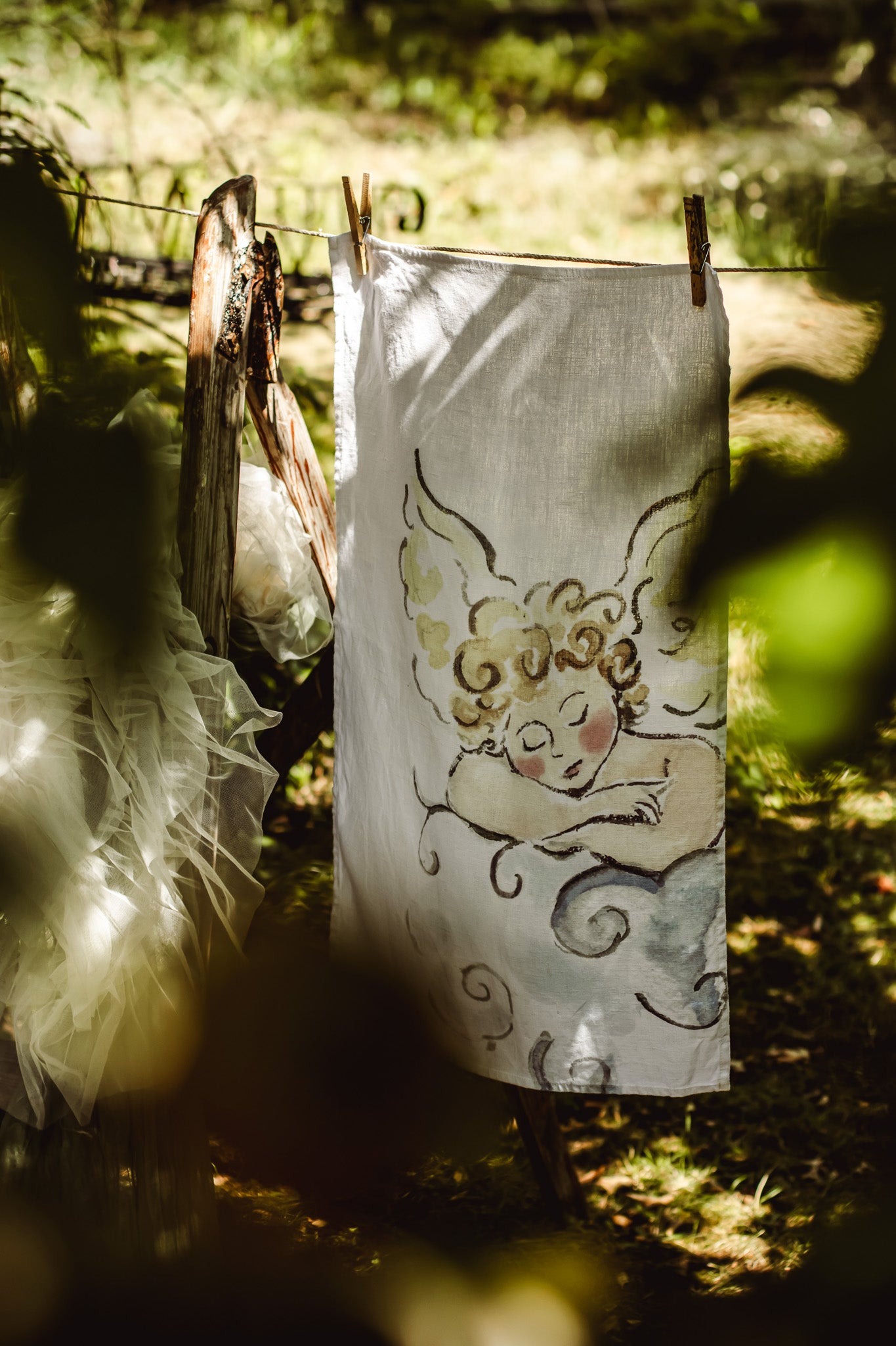 A white fabric tea towel or napkin with a hand-painted cherub design, hanging from a clothesline outdoors in a sunlit forest setting.