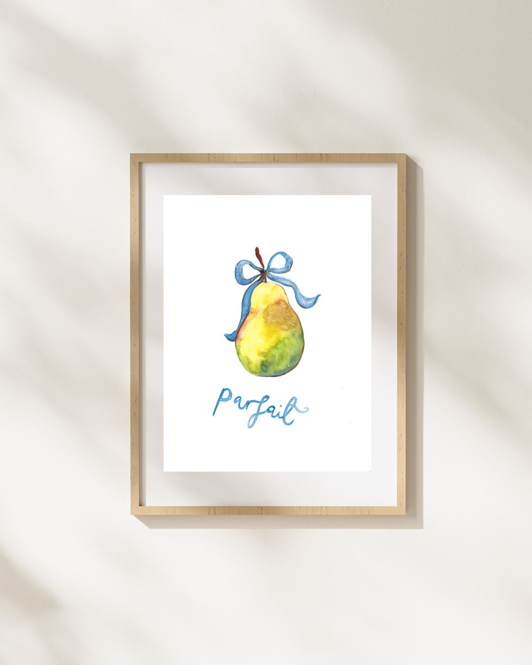 A framed watercolor art print featuring a green and yellow pear tied with a blue ribbon and the word 'perfect' in blue script, displayed on a warm off-white wall with soft shadow overlays.
