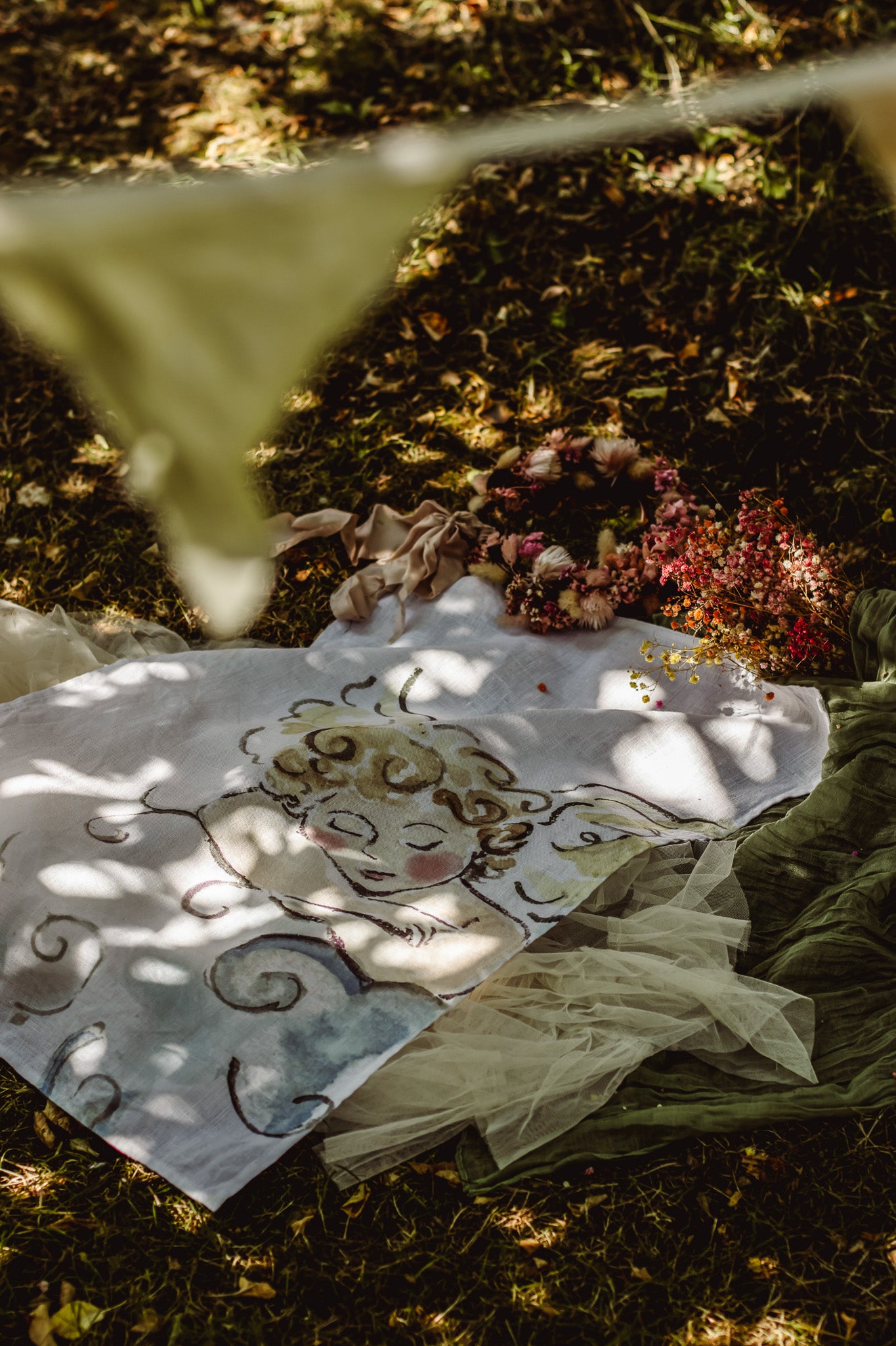 A white fabric blanket with a whimsical hand-drawn illustration of a sleeping angel, styled outdoors on grass with dappled sunlight and boho decor.