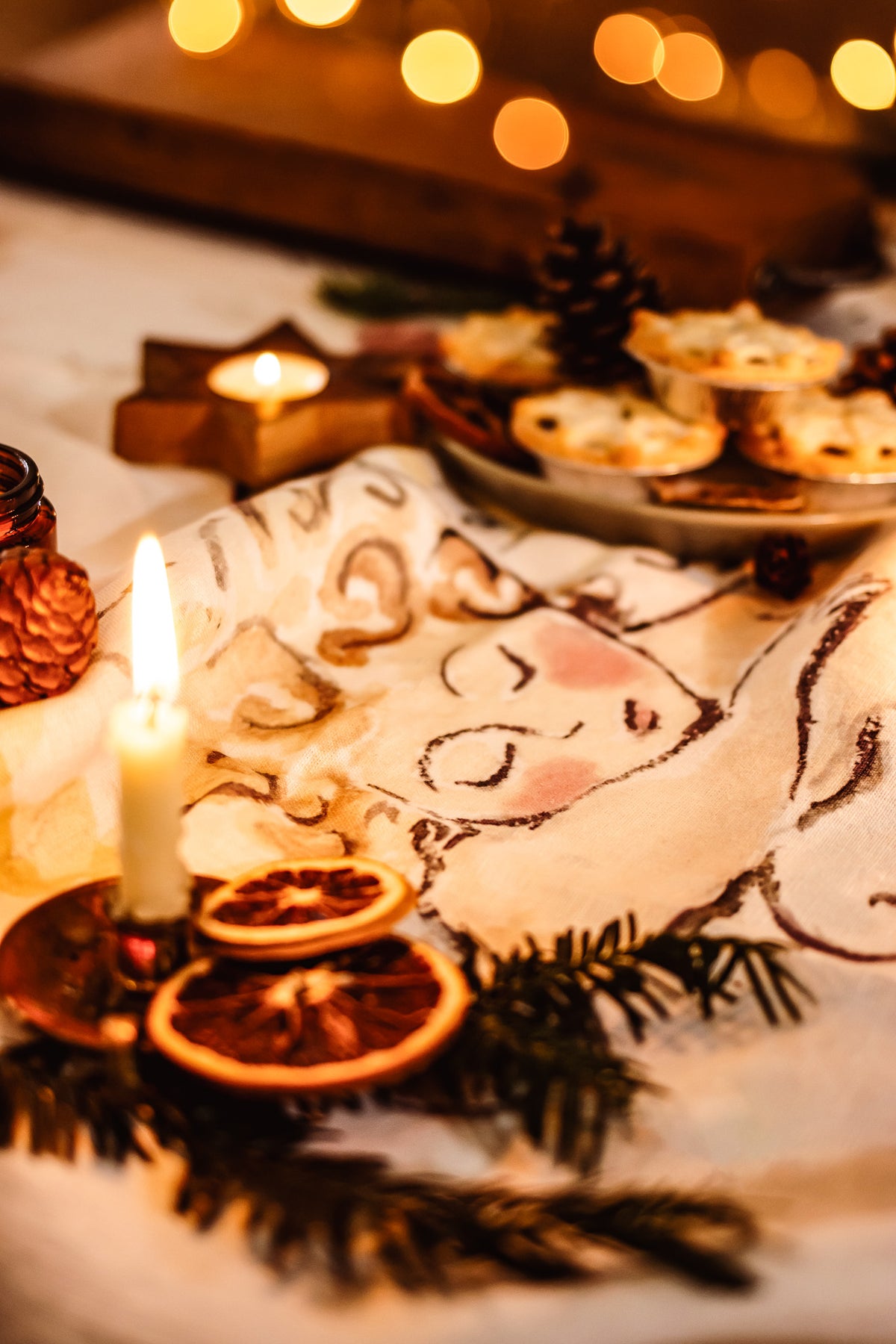 A festive Christmas table setting with a white tablecloth printed with a sleeping angel design, decorated with pine branches, dried orange slices, a lit candle, and a plate of mince pies under warm golden bokeh lights.