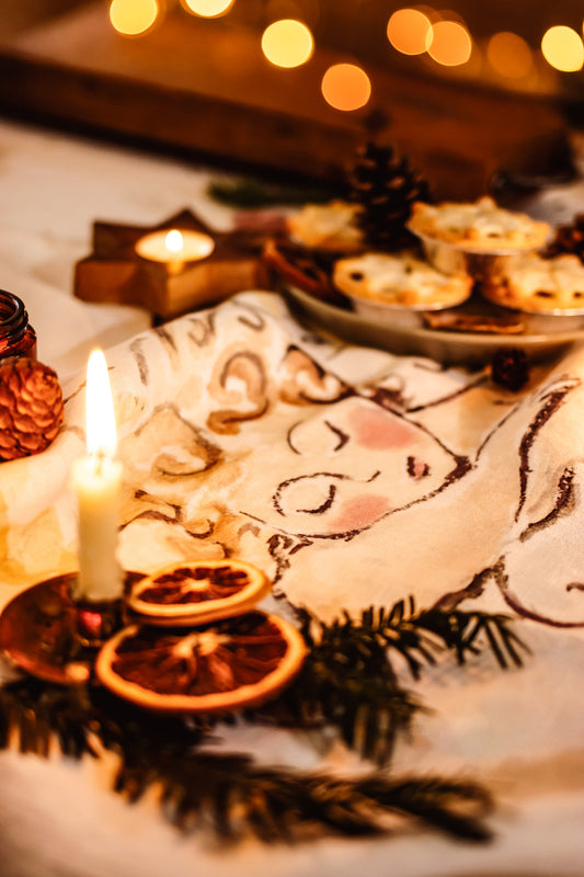 A festive Christmas table setting with a white tablecloth printed with a sleeping angel design, decorated with pine branches, dried orange slices, a lit candle, and a plate of mince pies under warm golden bokeh lights.