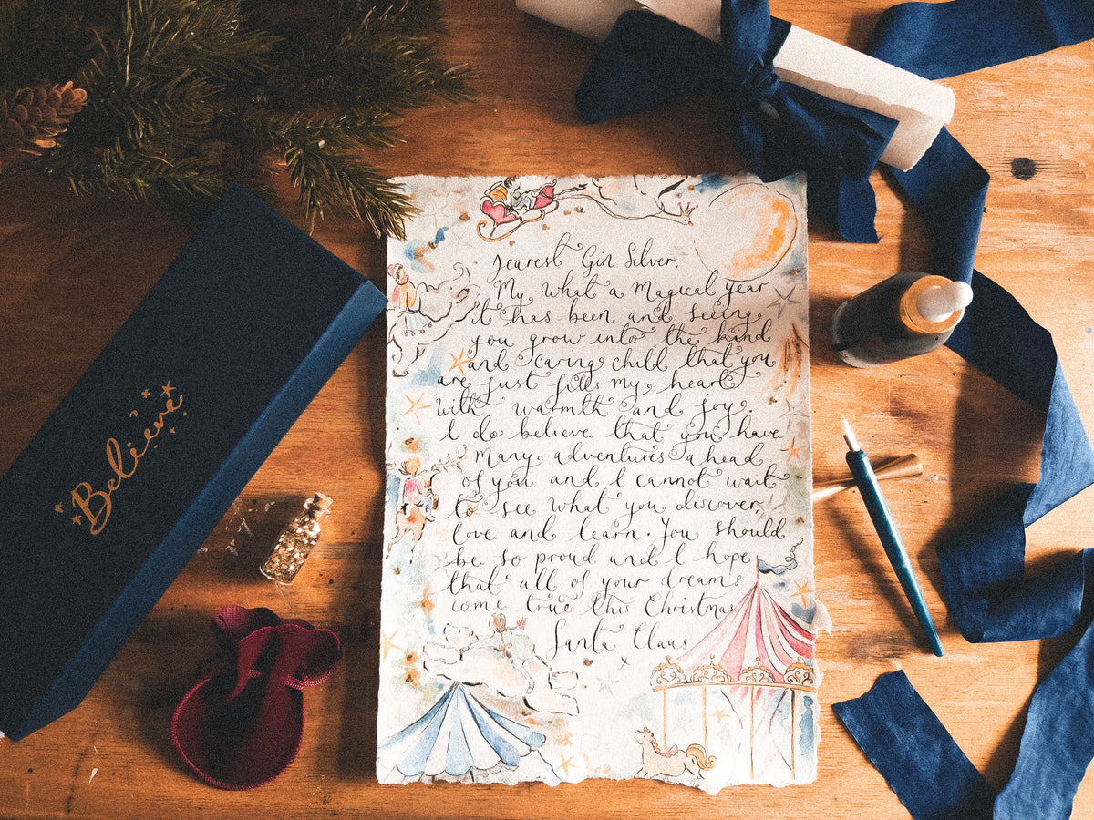 A beautifully styled flat-lay of a hand-lettered Christmas card on a wooden table, surrounded by a navy blue gift box with gold script, a small velvet pouch, a pen, and festive greenery.