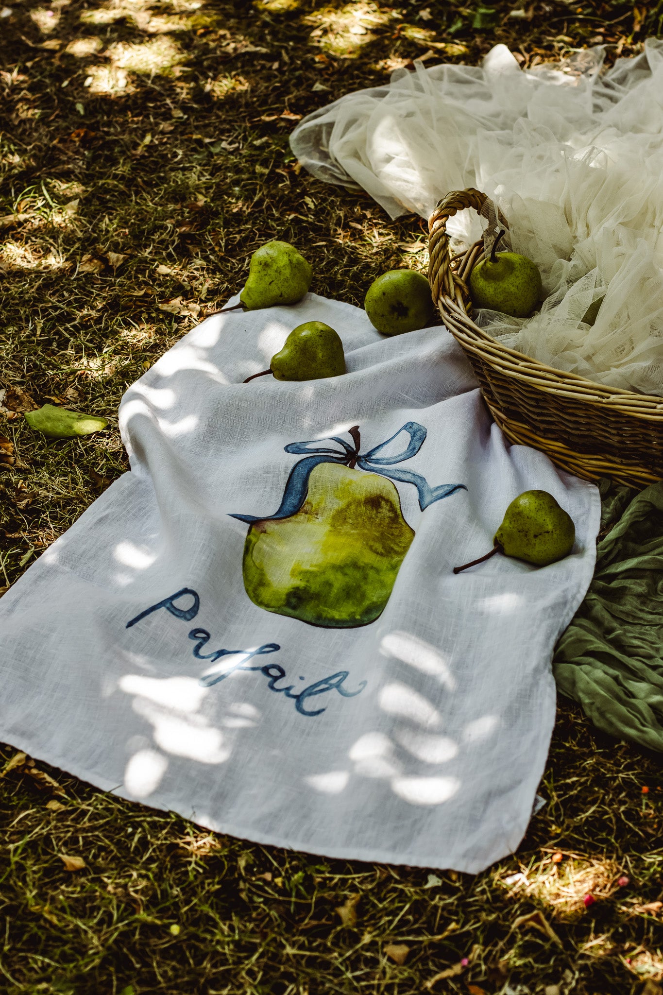 A white linen tea towel with a painted green pear and the word 'Pafal' in blue script, laid on grass outdoors with pears and a wicker basket nearby.