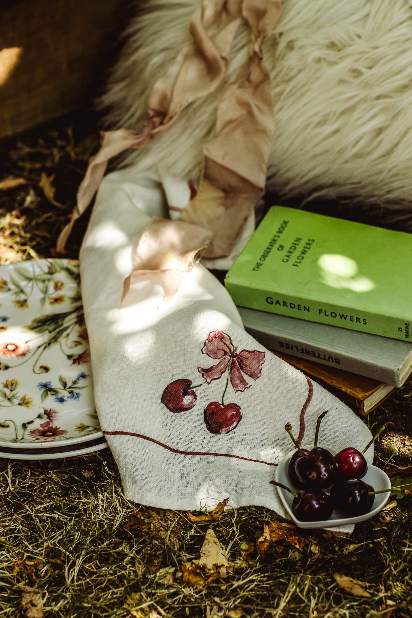 A cream linen napkin with embroidered cherries and a bow, styled on a rustic wooden surface with a green 'Garden Flowers' book, a ceramic plate, and a small bowl of cherries.