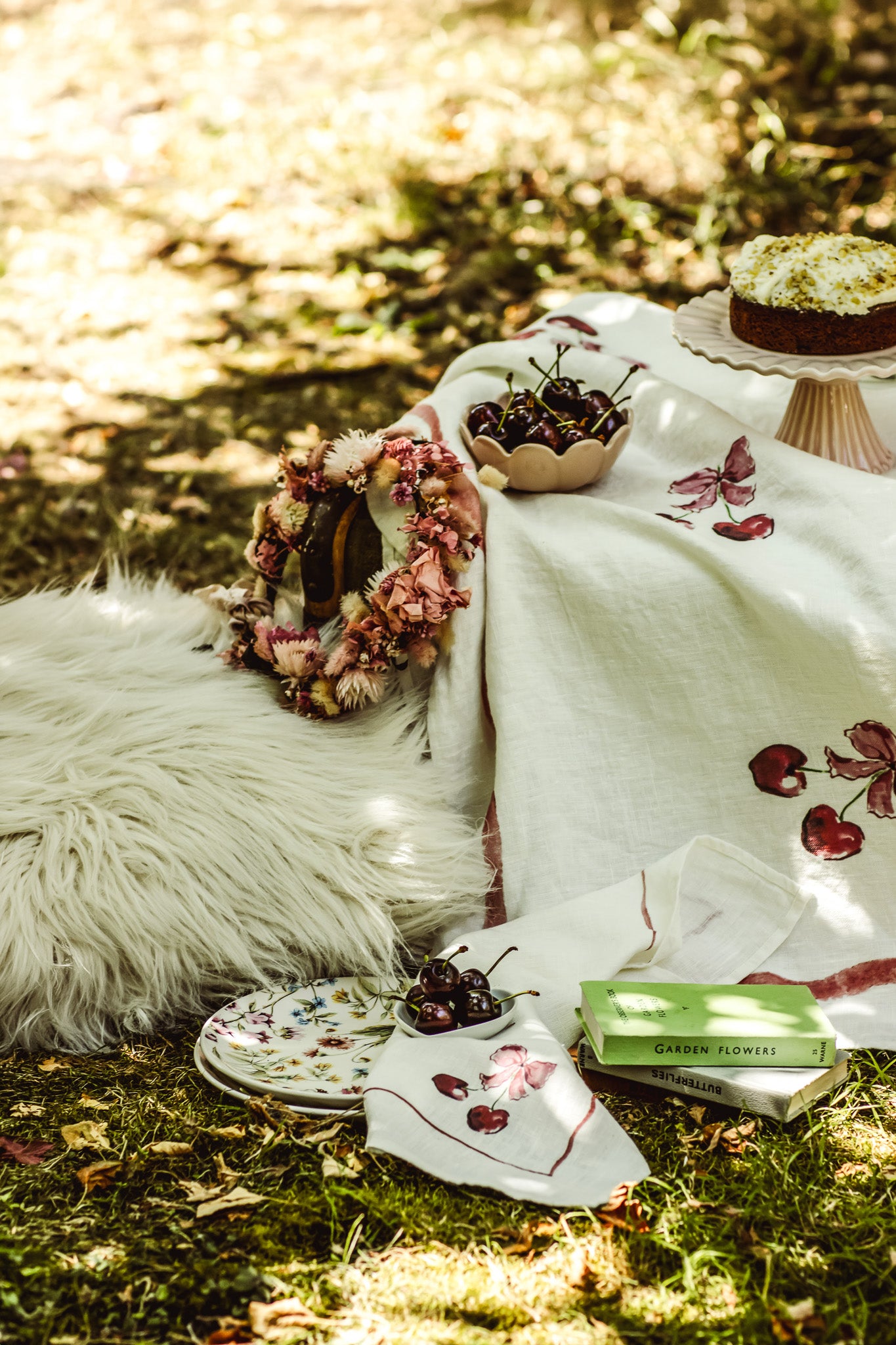 A styled picnic scene outdoors on grass with dappled sunlight, featuring a white embroidered blanket with red floral motifs, a white faux-fur cushion, a floral wreath, plates, cherries, and a green book.