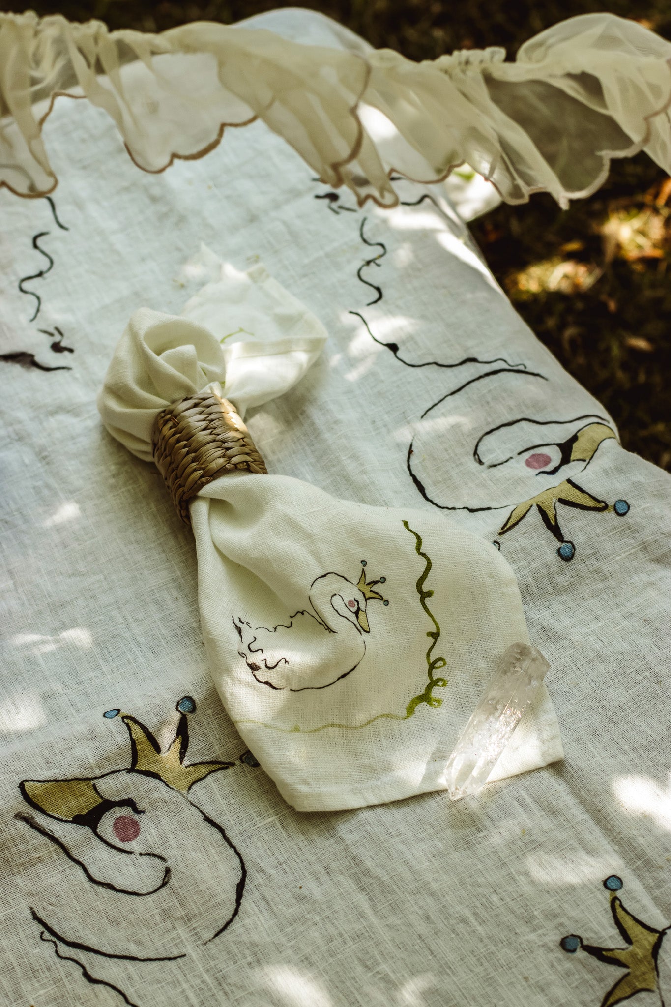 A cream-colored linen napkin with embroidered swan designs and a wicker band, resting on a matching patterned linen tablecloth outdoors under dappled sunlight.