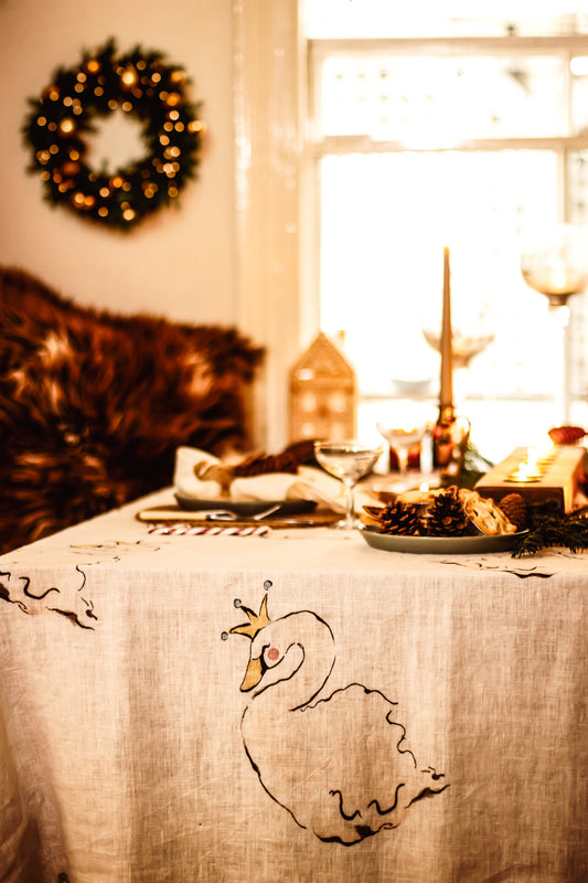 A beige linen tablecloth with a hand-drawn black swan design and a small gold crown, styled on a dining table in a warm, cozy room with a window and holiday wreath in the background.
