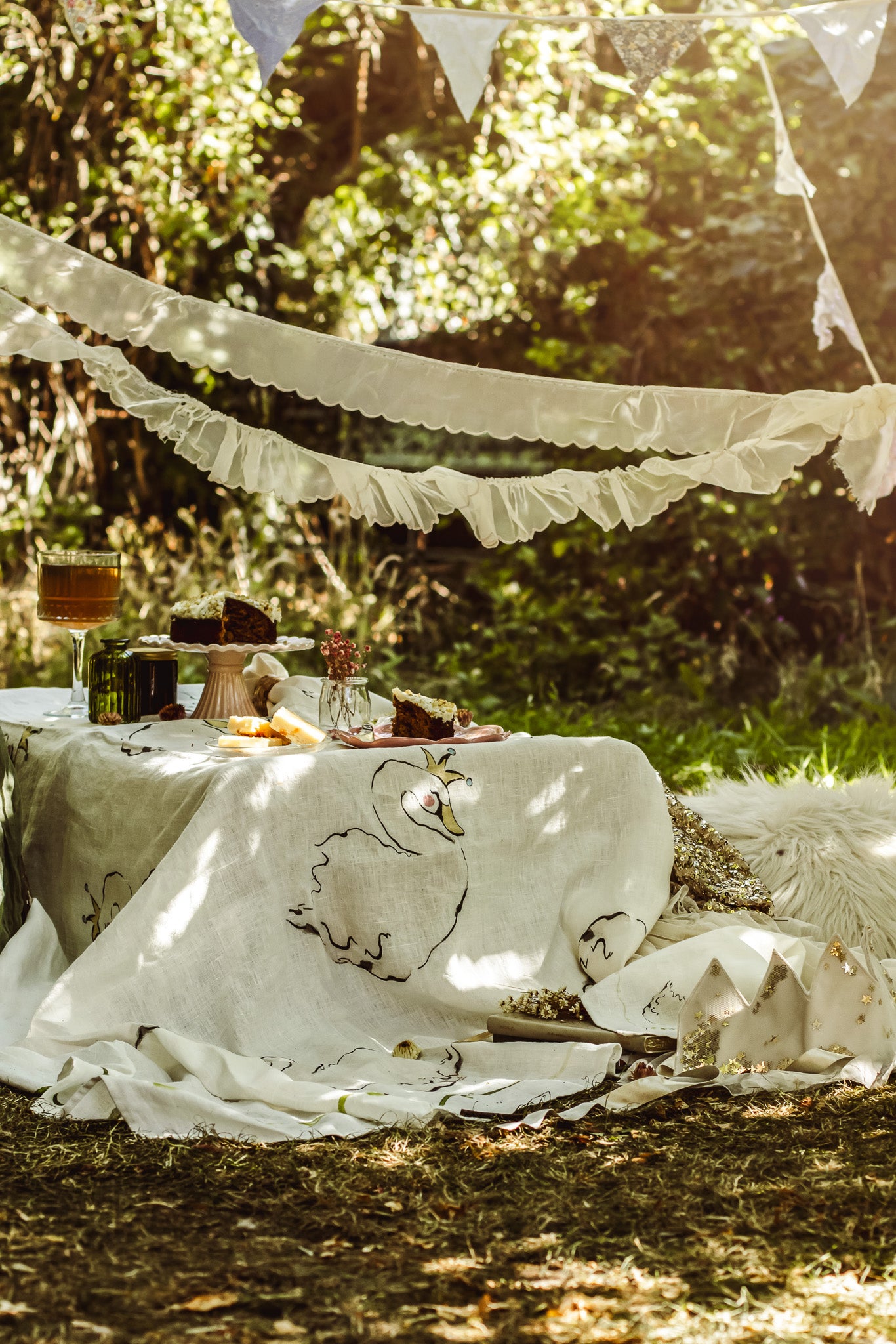 A rustic outdoor picnic setup with a cream-colored tablecloth featuring a whimsical duck illustration, draped over a table with food, set against a backdrop of lush green trees with dappled sunlight.