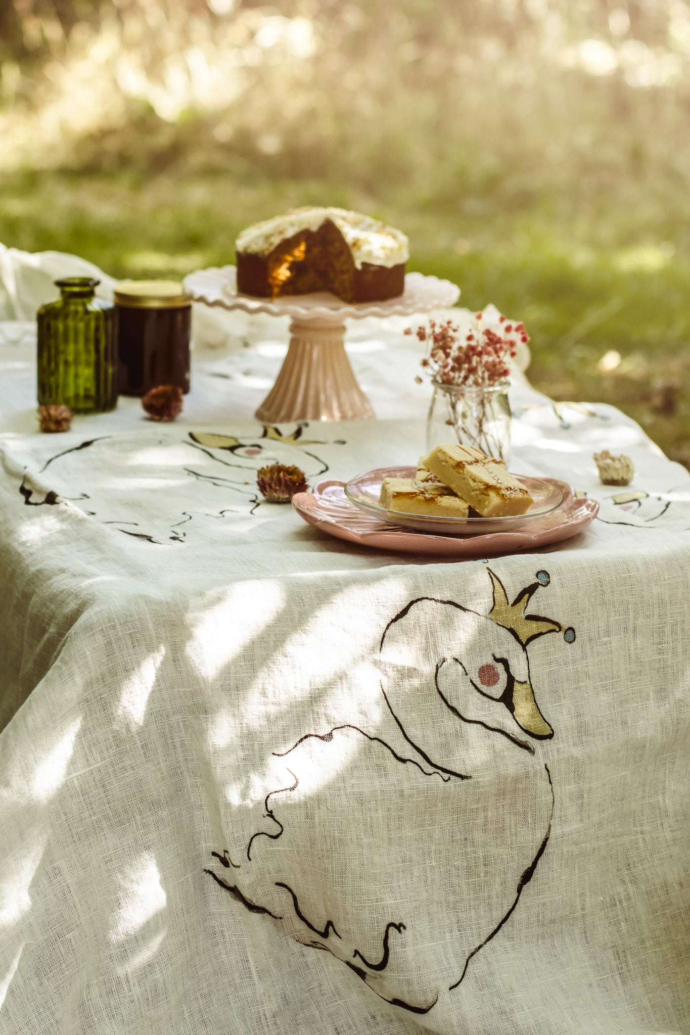 A white linen tablecloth with a whimsical black embroidered bird design, styled outdoors on a table with a slice of cake, plates, and small glass bottles in a sunlit garden setting.