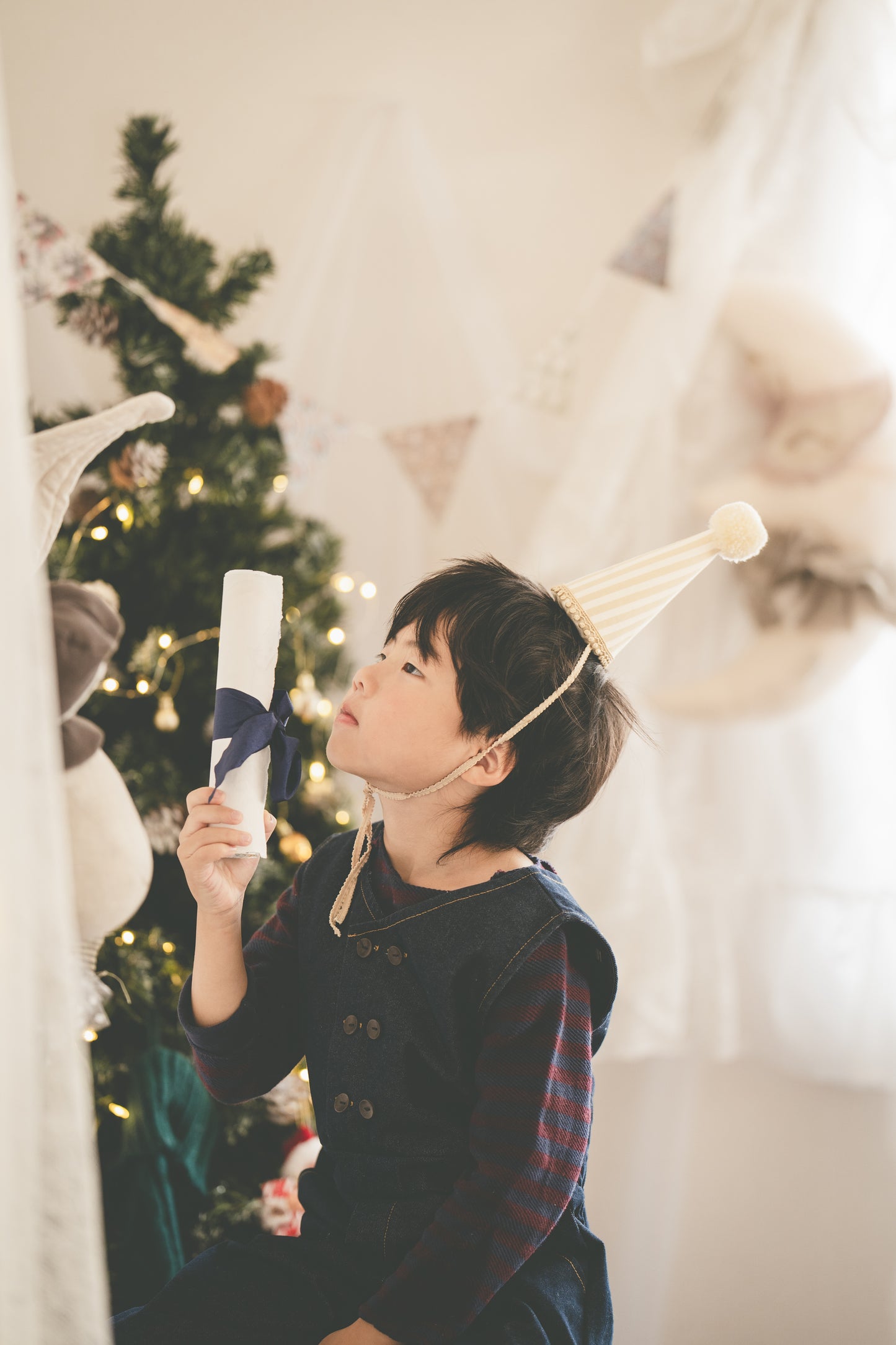 A young boy wearing a dark navy pinafore vest over a red plaid shirt, holding a rolled white paper certificate or scroll. He is wearing a cream party hat and looking upwards in a softly lit room with a decorated Christmas tree in the background.