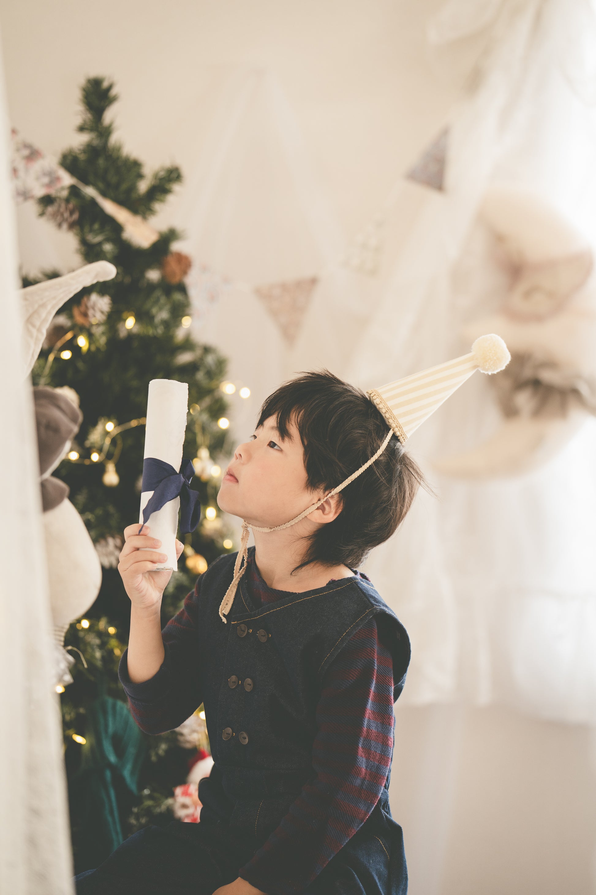 A young boy wearing a dark navy pinafore vest over a red plaid shirt, holding a rolled white paper certificate or scroll. He is wearing a cream party hat and looking upwards in a softly lit room with a decorated Christmas tree in the background.