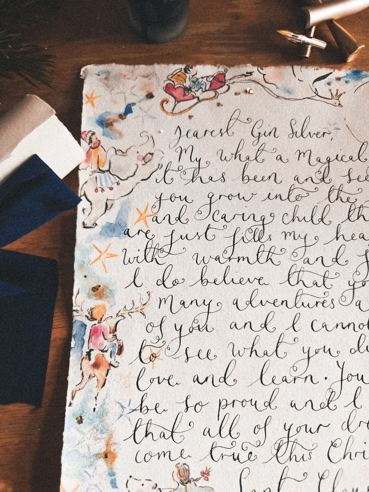 A close-up of a decorative, hand-lettered paper card or print featuring cursive text and whimsical watercolor illustrations of children and stars, styled on a dark wooden surface with a navy napkin and gold pen visible around it.