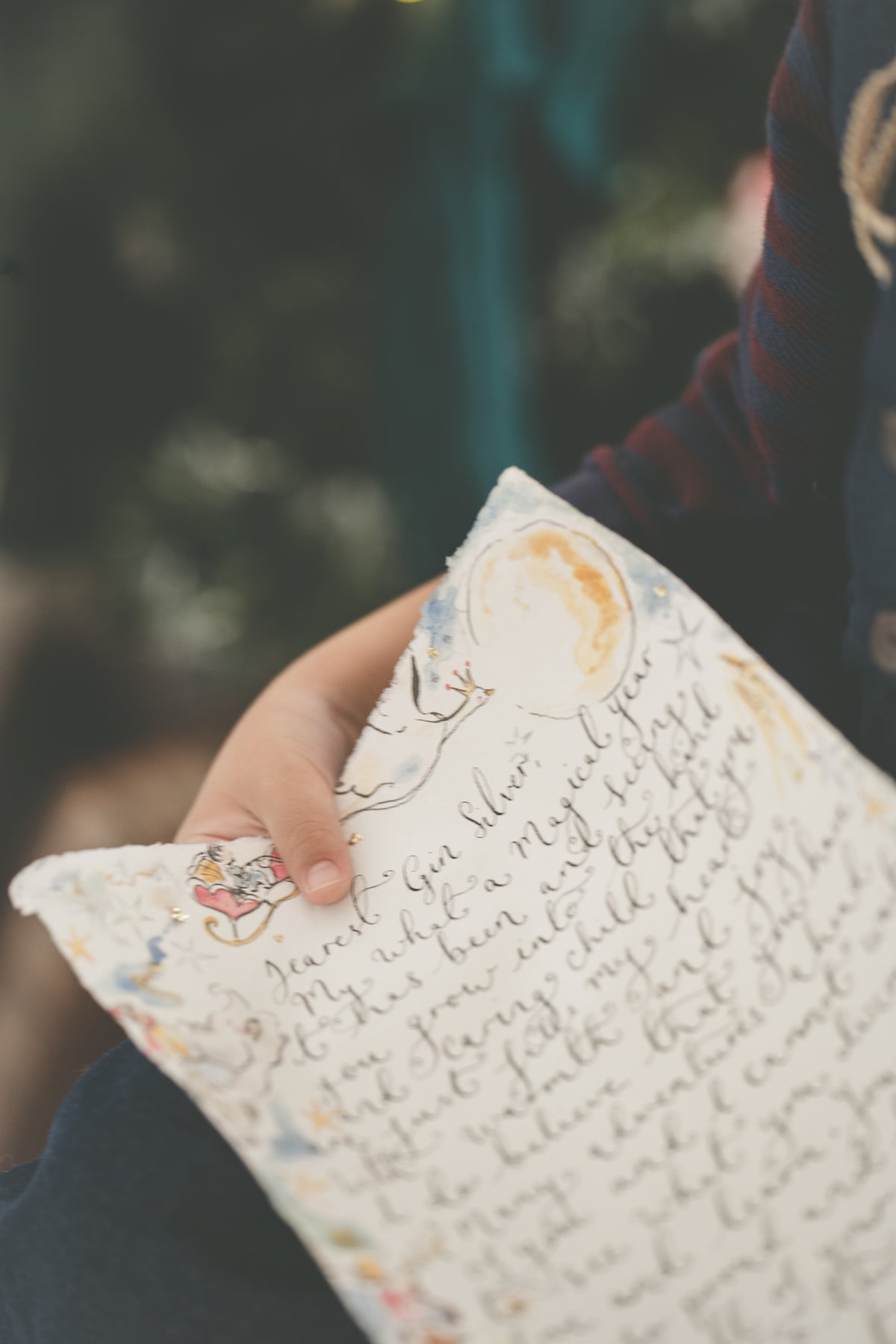A close-up lifestyle shot of a hand holding a letter. The pillow is off-white with a watercolor illustration of a bird and moon, and handwritten cursive text.