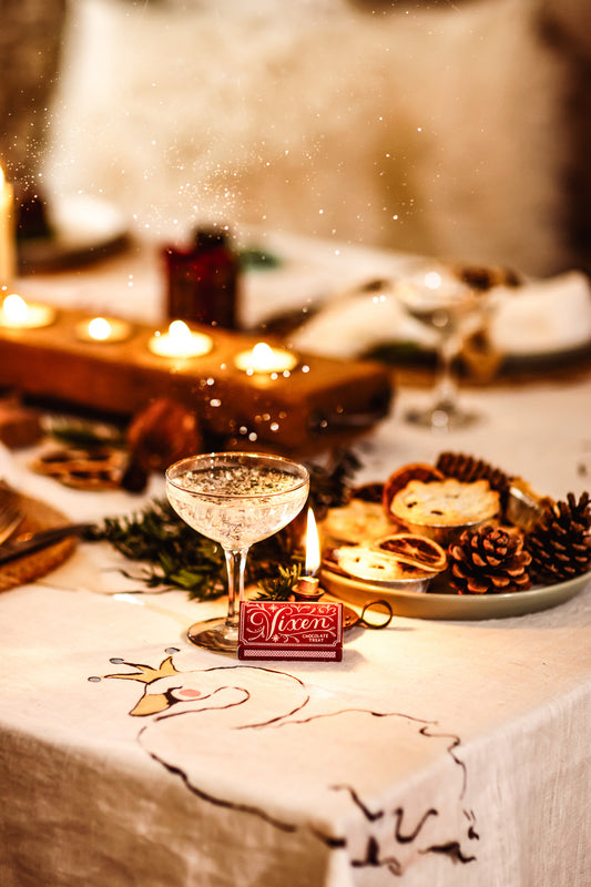A festive holiday table setting with a lit champagne flute, pinecones, candles, and a small red sign reading 'Merry' on a warm, softly blurred background.