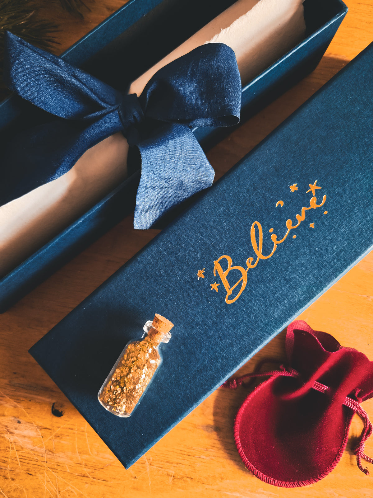 A dark blue velvet gift box with gold script reading 'Believe' lies on a wooden surface next to an open box tied with a navy ribbon, a small glass vial with dried botanicals, and a red velvet drawstring pouch.