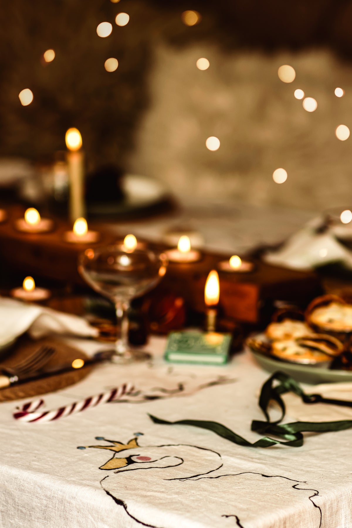 A cozy, warmly lit table setting with a white embroidered tablecloth featuring a cartoon snowman and the word 'Winter', surrounded by lit candles, a wine glass, and soft bokeh lights in the background.