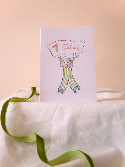 Greeting card with illustration of a child holding a '1 today' sign on a white surface with a green ribbon.