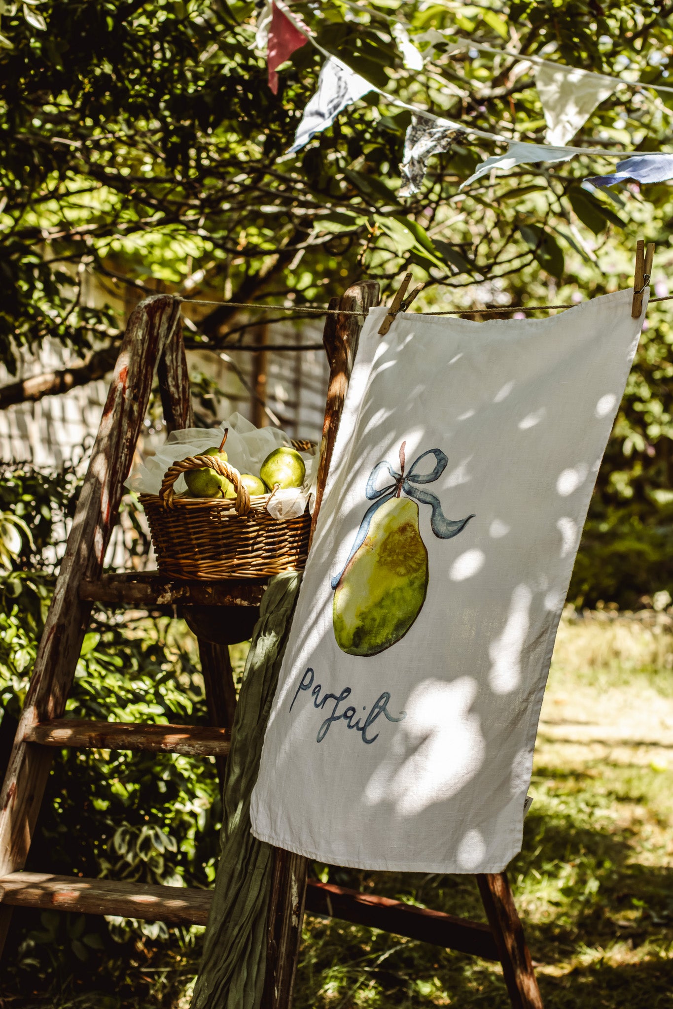 A white tea towel with a watercolor illustration of a green pear and the word 'perfume' in script, hanging outdoors on a rustic wooden ladder in a garden setting.