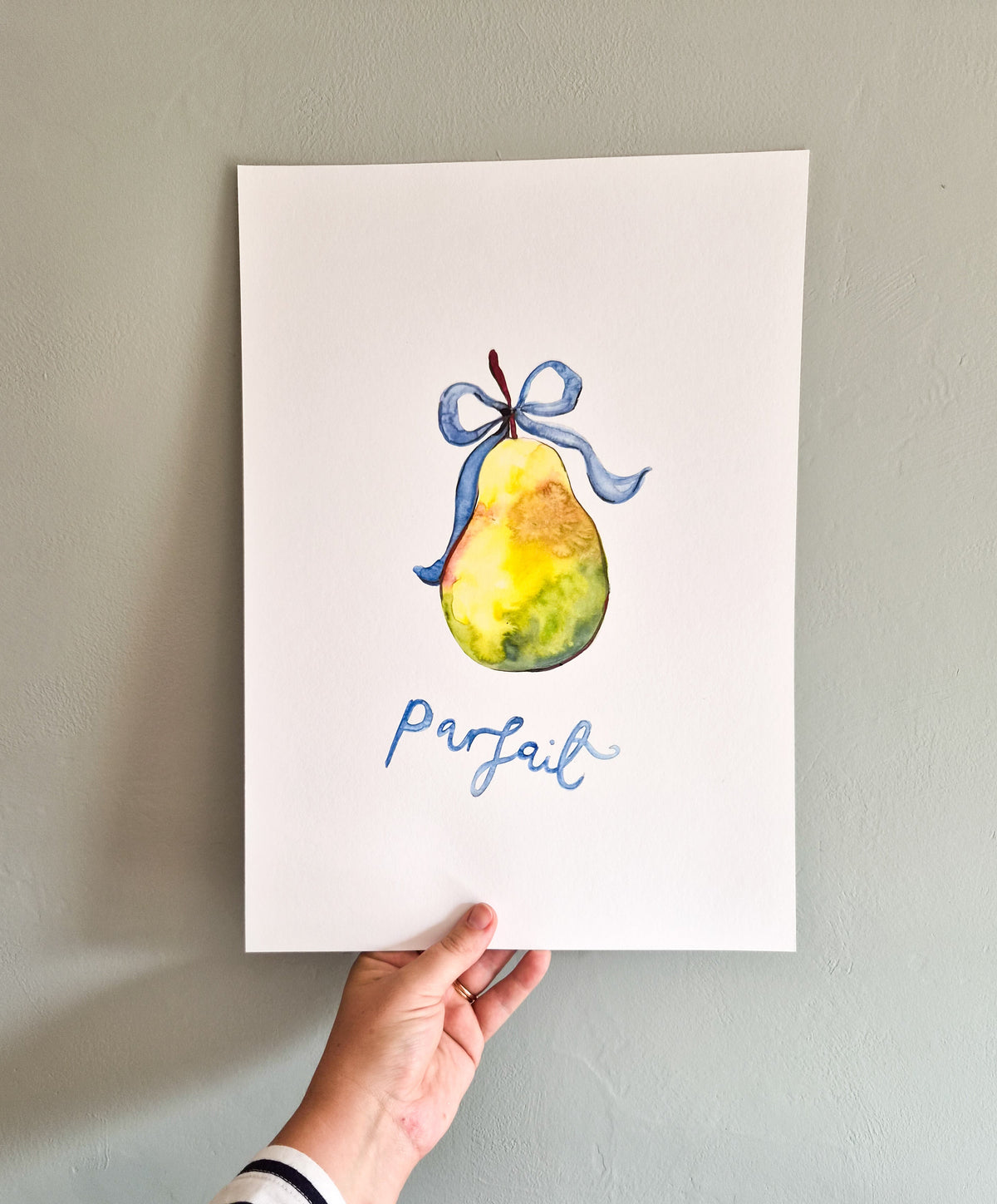 A hand holds a white art print featuring a watercolor illustration of a yellow pear with a blue ribbon and the word 'parfait' in blue script, against a light gray wall.