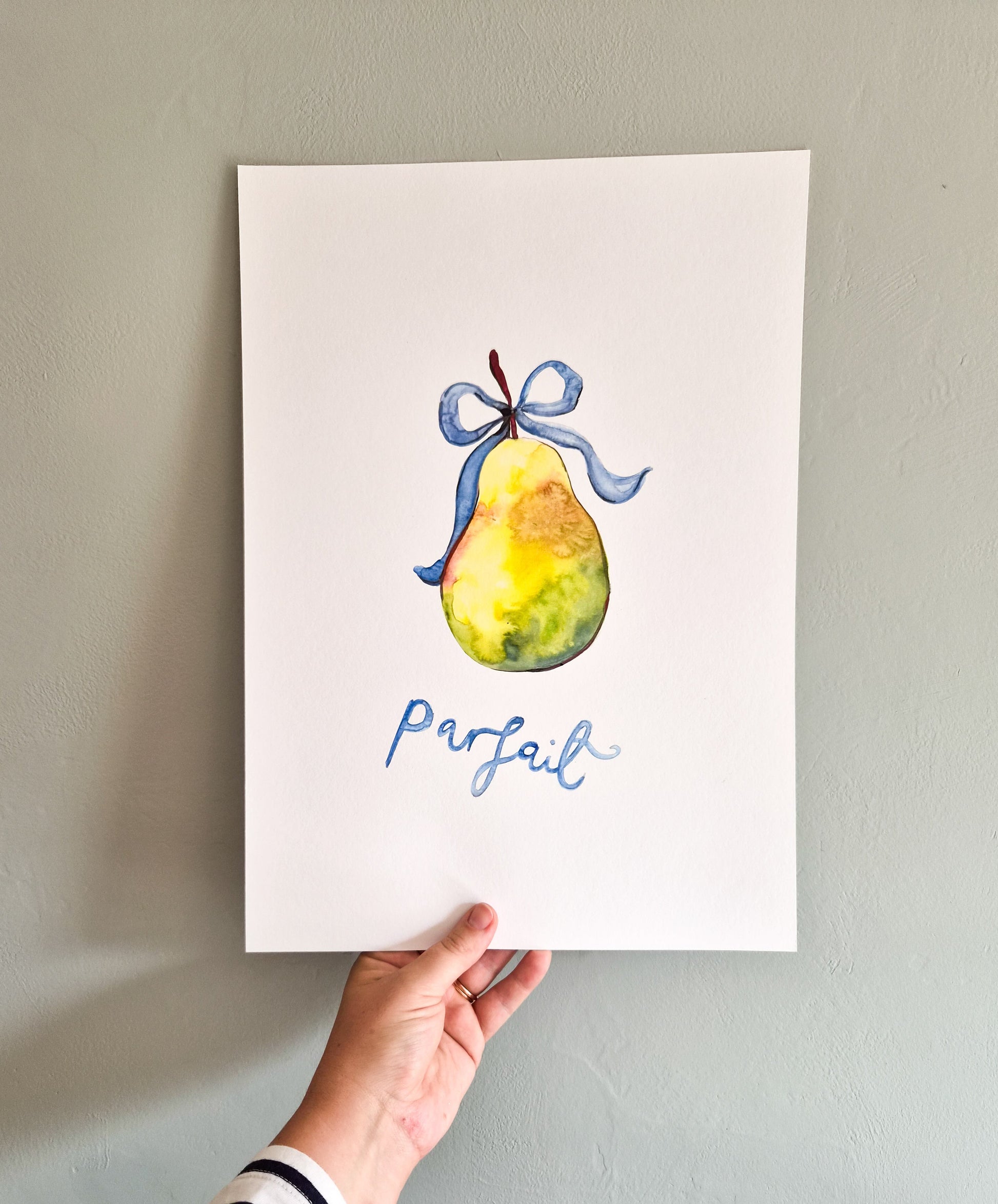 A hand holds a white art print featuring a watercolor illustration of a yellow pear with a blue ribbon and the word 'parfait' in blue script, against a light gray wall.