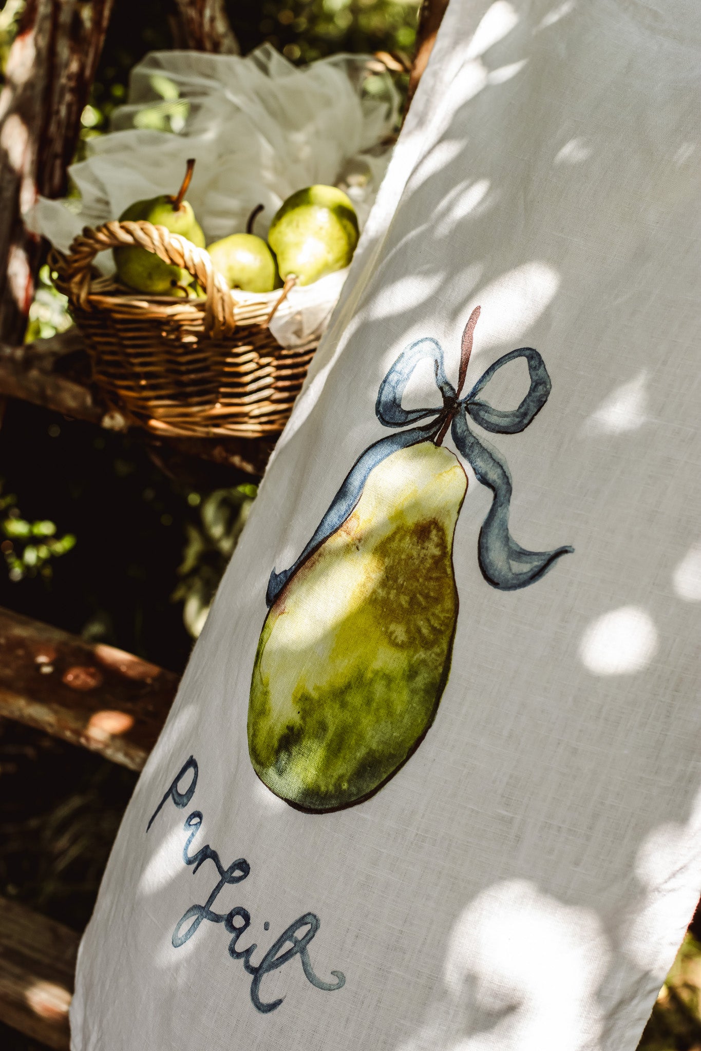 A close-up of a white linen fabric, likely a napkin or table linen, featuring a painted green pear tied with a blue ribbon and the word 'Perfait' written in black script. The background shows a rustic wicker basket filled with green pears in a sun-dappled outdoor setting.