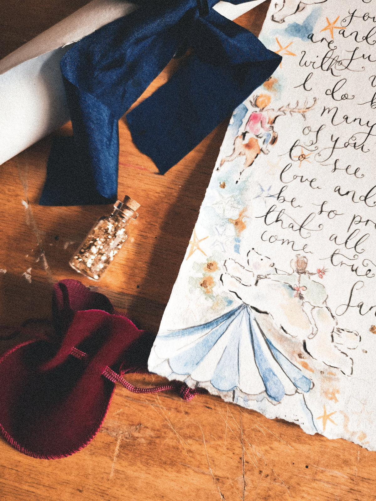 A styled flat-lay of a handmade paper bookmark or art print featuring watercolor illustrations of cherubs and cursive writing, accompanied by a navy blue ribbon, a small glass bottle with glitter, and a burgundy velvet pouch on a wooden surface.