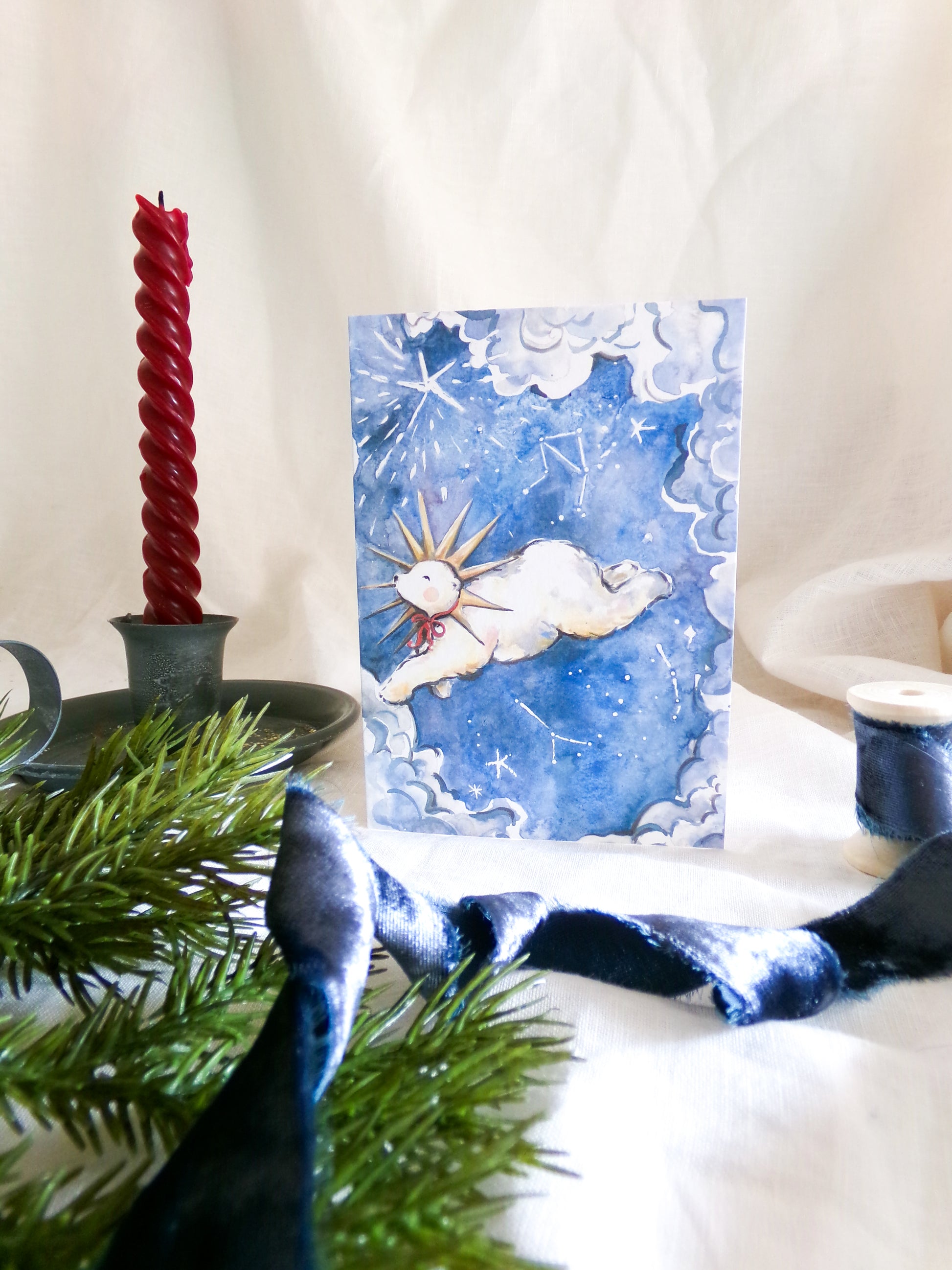 A square art print or card featuring a whimsical illustration of a white polar bear with a gold crown on a blue watercolor background, styled with a red twisted candle, green pine branches, and dark blue ribbon on a white fabric backdrop.