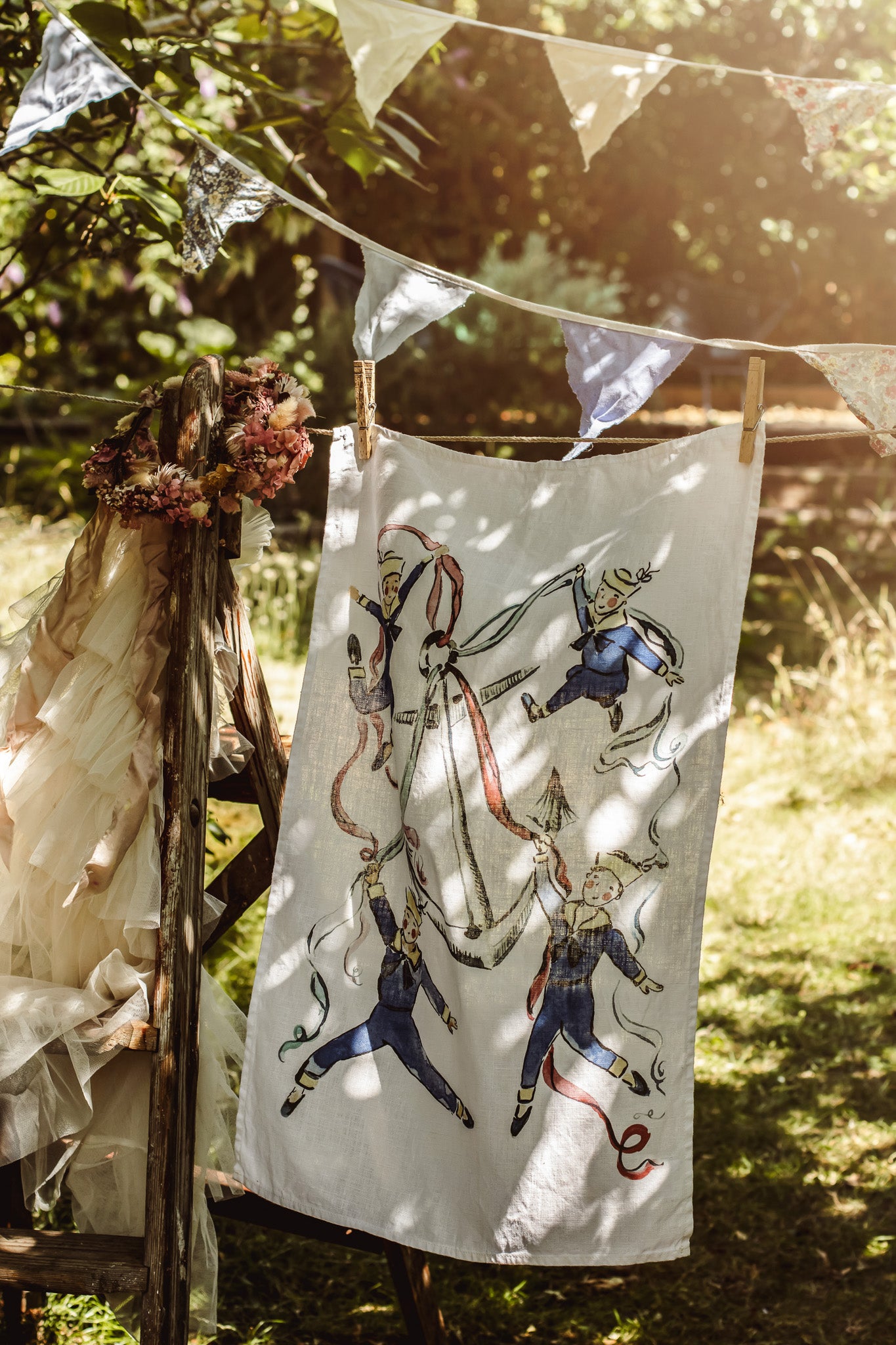 A white fabric tea towel or napkin printed with a vintage illustration of soldiers in blue and red uniforms, hanging from a wooden stand outdoors on a string with bunting flags.