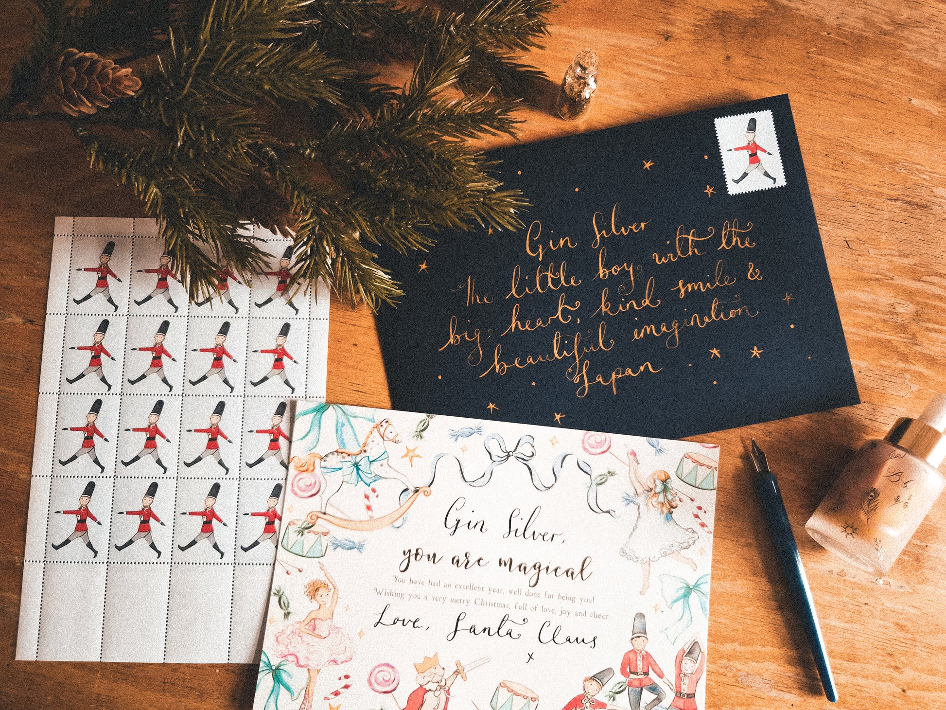 A styled flat-lay of Christmas-themed stationery on a wooden surface, including a black card with gold lettering, a sheet of postage stamps, a colorful illustrated card, pine branches, a pen, and a small bottle.