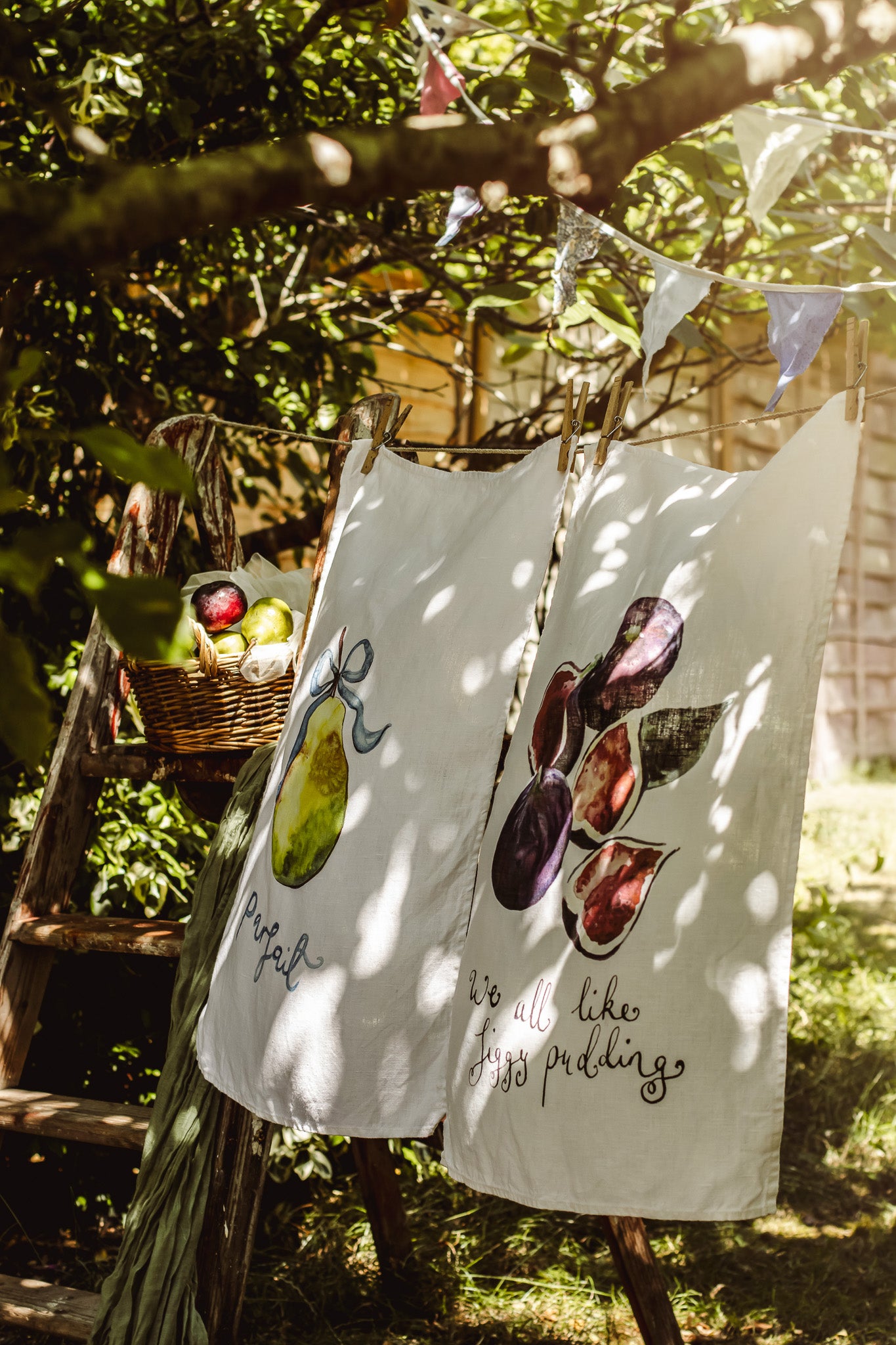 Two white tea towels with painted fruit illustrations and cursive text hanging from a wooden ladder outdoors in a garden setting.
