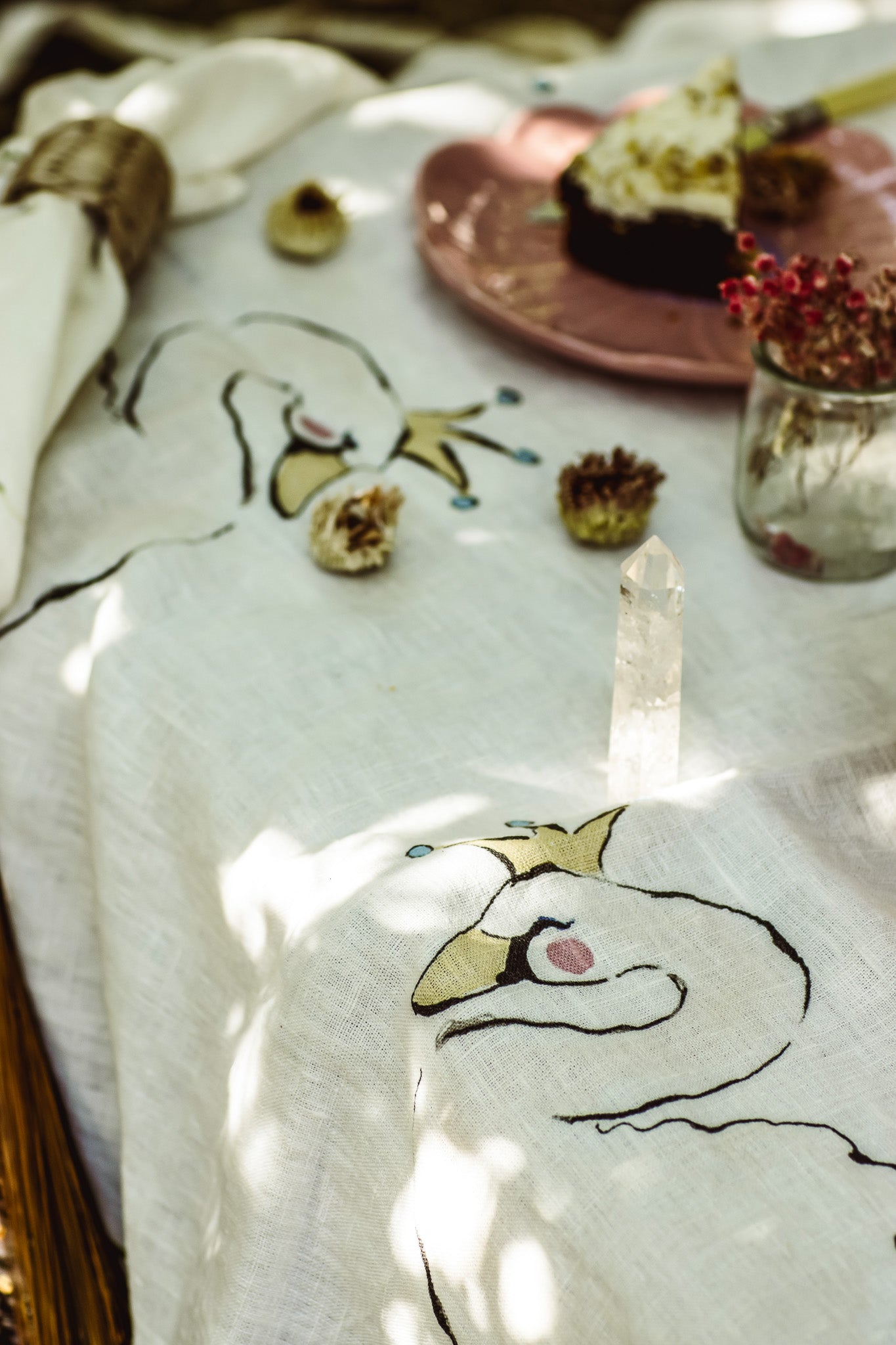 A white linen tablecloth featuring embroidered swan designs with gold crowns and pink accents, styled with a crystal point, dried flowers, and a slice of cake in a rustic setting.
