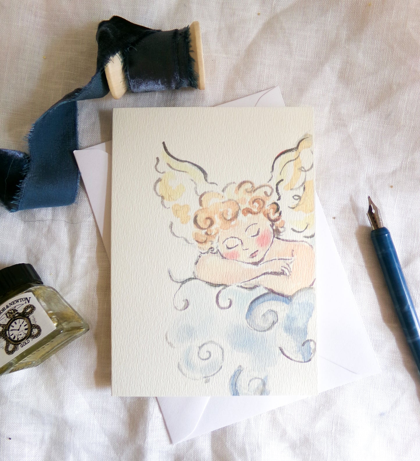 A square art print or card featuring a watercolor illustration of a sleeping angel with curly golden hair and blue clouds, styled on a white fabric surface with a blue ribbon, ink bottle, and fountain pen nearby.