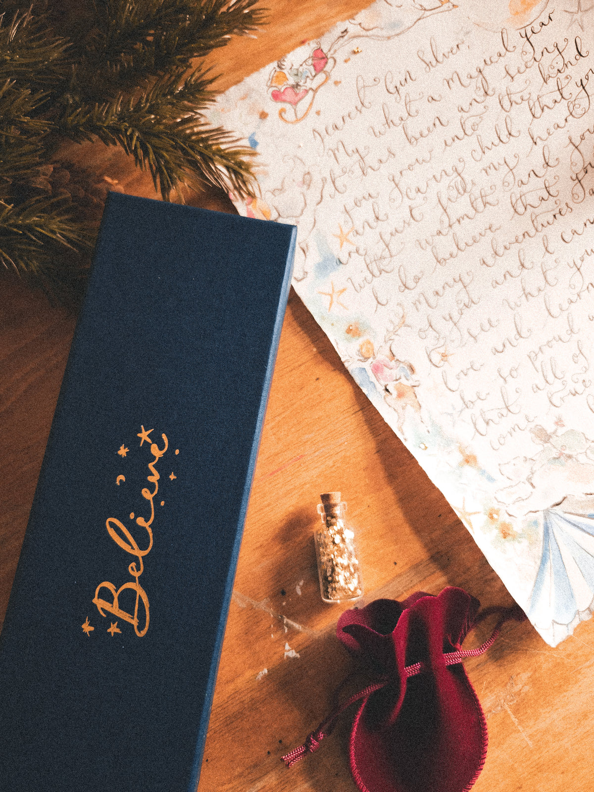 A dark navy blue gift box with gold foil 'Believe' script, styled on a wooden surface next to a handwritten letter, a small glass vial, and a red velvet pouch.