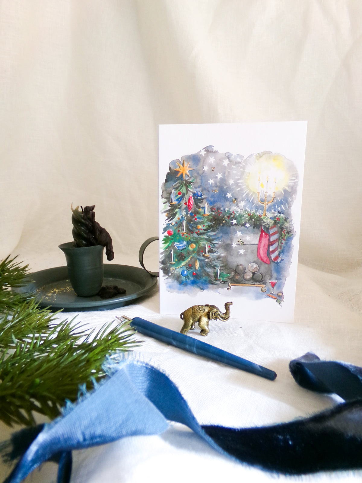 A vertical greeting card with a watercolor Christmas scene featuring a decorated tree, a glowing star, and snowflakes, styled on a white fabric backdrop with a blue pen, a small elephant figurine, and greenery.