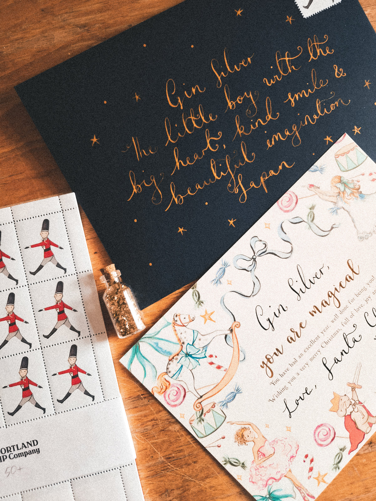 A styled flat-lay of a Gin Silver postage stamp sheet and matching illustrated postcard on a wooden surface, with a small bottle of gold glitter nearby.