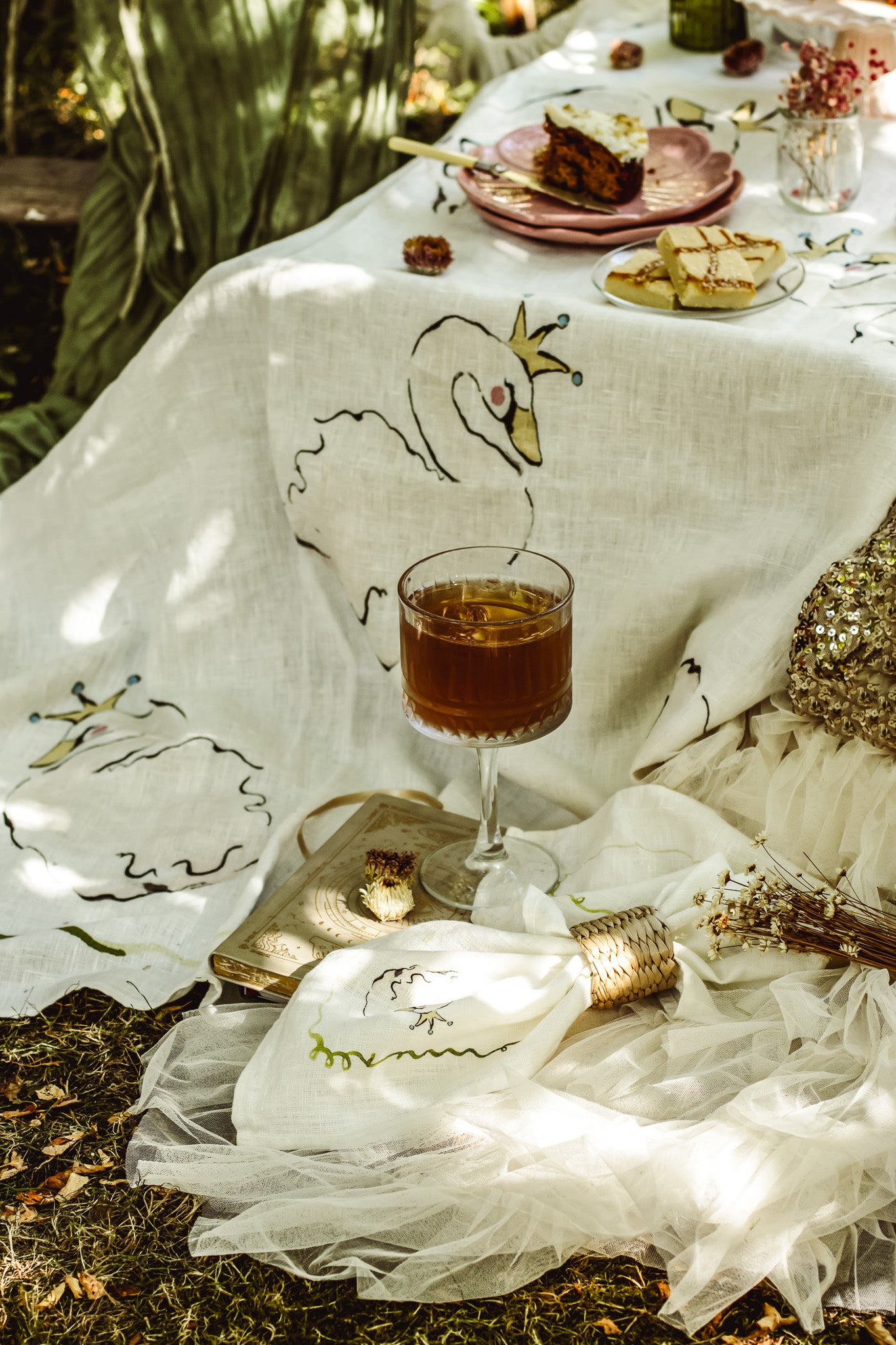 A bohemian-style outdoor picnic setup featuring a white linen tablecloth with embroidered swans, a glass of iced tea, and rustic plates on a forest floor.