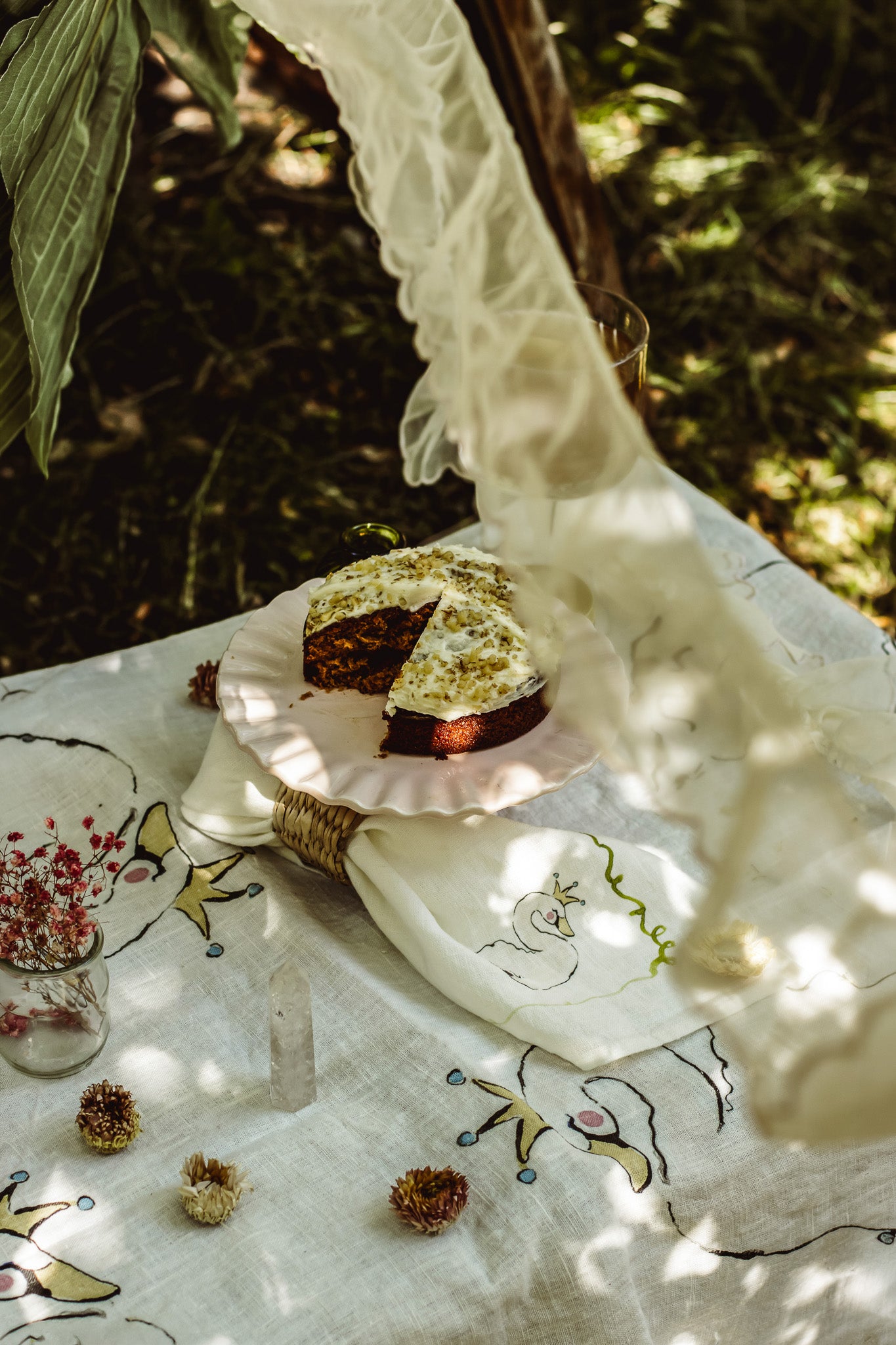 A rustic outdoor table setting with a slice of chocolate cake on a pink plate, draped with a cream lace cloth over a white embroidered linen tablecloth, with dried flowers and greenery in the background.