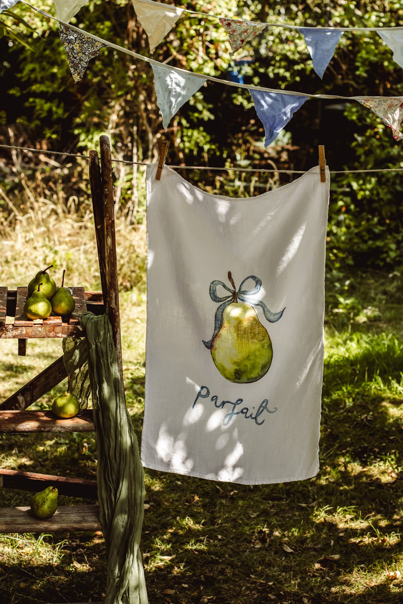 A white kitchen towel with a painted green pear and the word 'Parfait' hanging on a clothesline outdoors, with bunting and a rustic wooden bench in a sunlit garden setting.