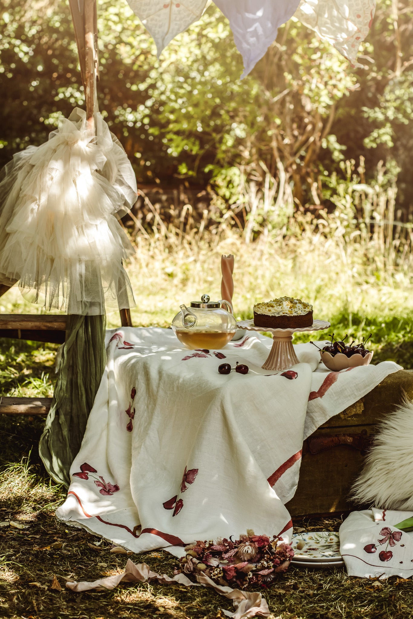 A styled outdoor picnic scene in a sunlit forest clearing, featuring a white embroidered blanket draped over a wooden bench, with a teapot, cake on a stand, and rustic natural surroundings.