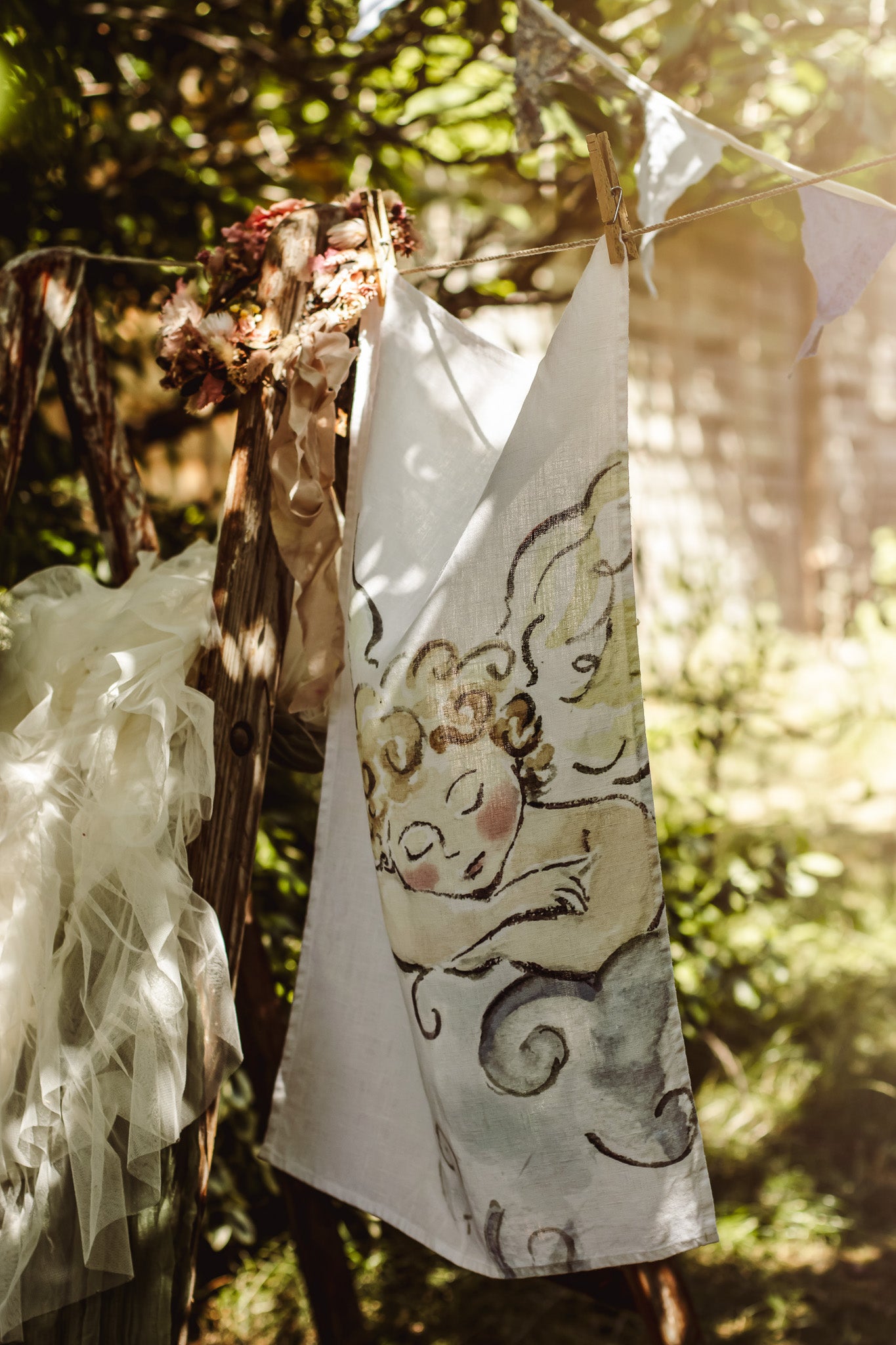 A white fabric napkin or tea towel with a watercolor illustration of a sleeping angel, hanging from a string outdoors with wooden clothespins against a sunlit, blurred garden background.
