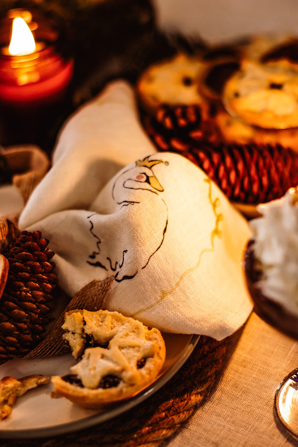 A close-up of a rustic holiday table setting featuring a white linen napkin embroidered with a robin design, resting on a plate with a half-eaten mince pie. The scene is decorated with pinecones, a burning red candle in the background, and more pies on a table runner.