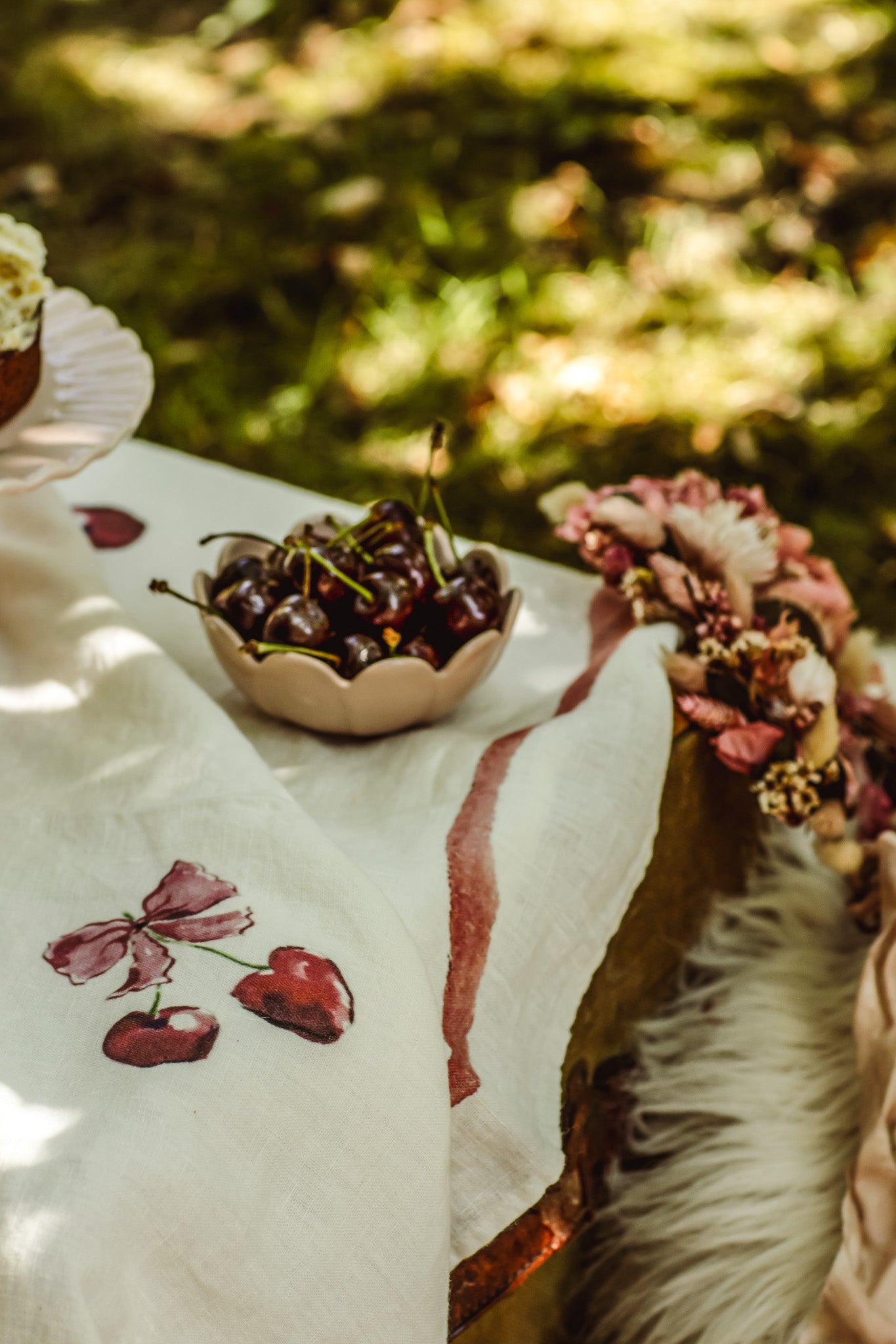 A close-up of a white embroidered tablecloth with cherry motifs, featuring a small bowl of fresh cherries and a floral arrangement in an outdoor garden setting.