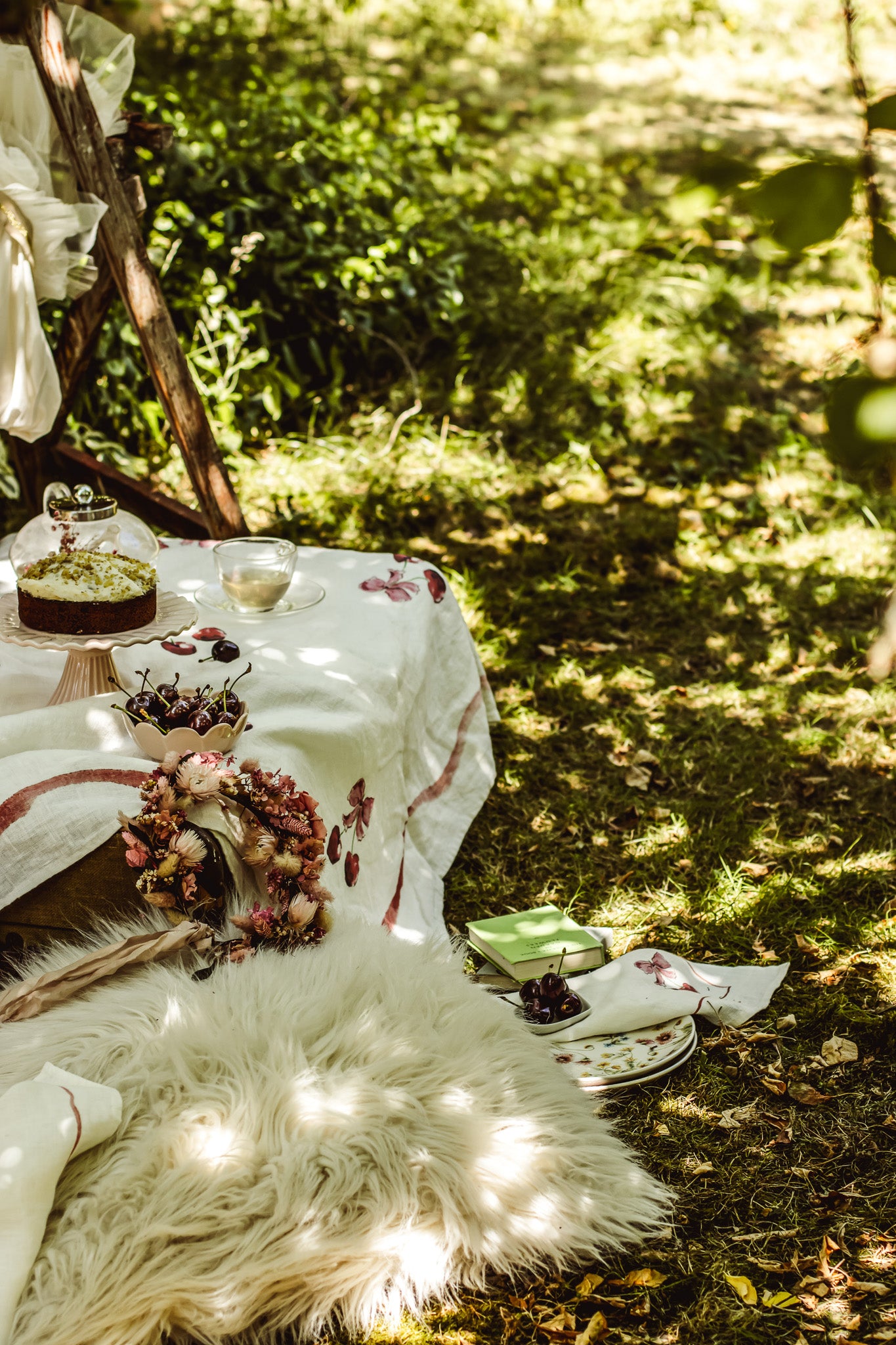 Outdoor picnic setup in a sun-dappled forest clearing, with a white tablecloth, plates, a small cake, flowers, and a fluffy cream rug on the grass.