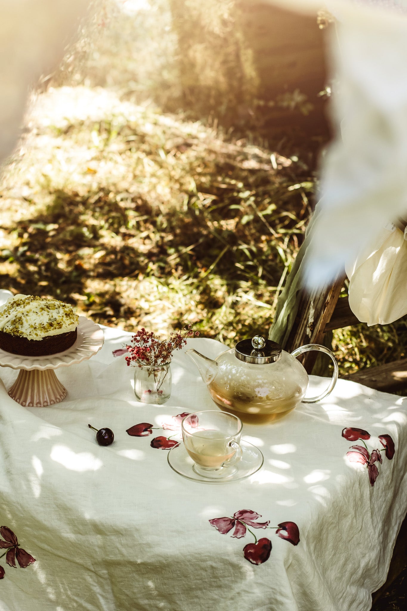 A styled outdoor tea setup on a white embroidered tablecloth with a glass teapot, teacup, flowers, and a cake, set against a sun-dappled grassy background.