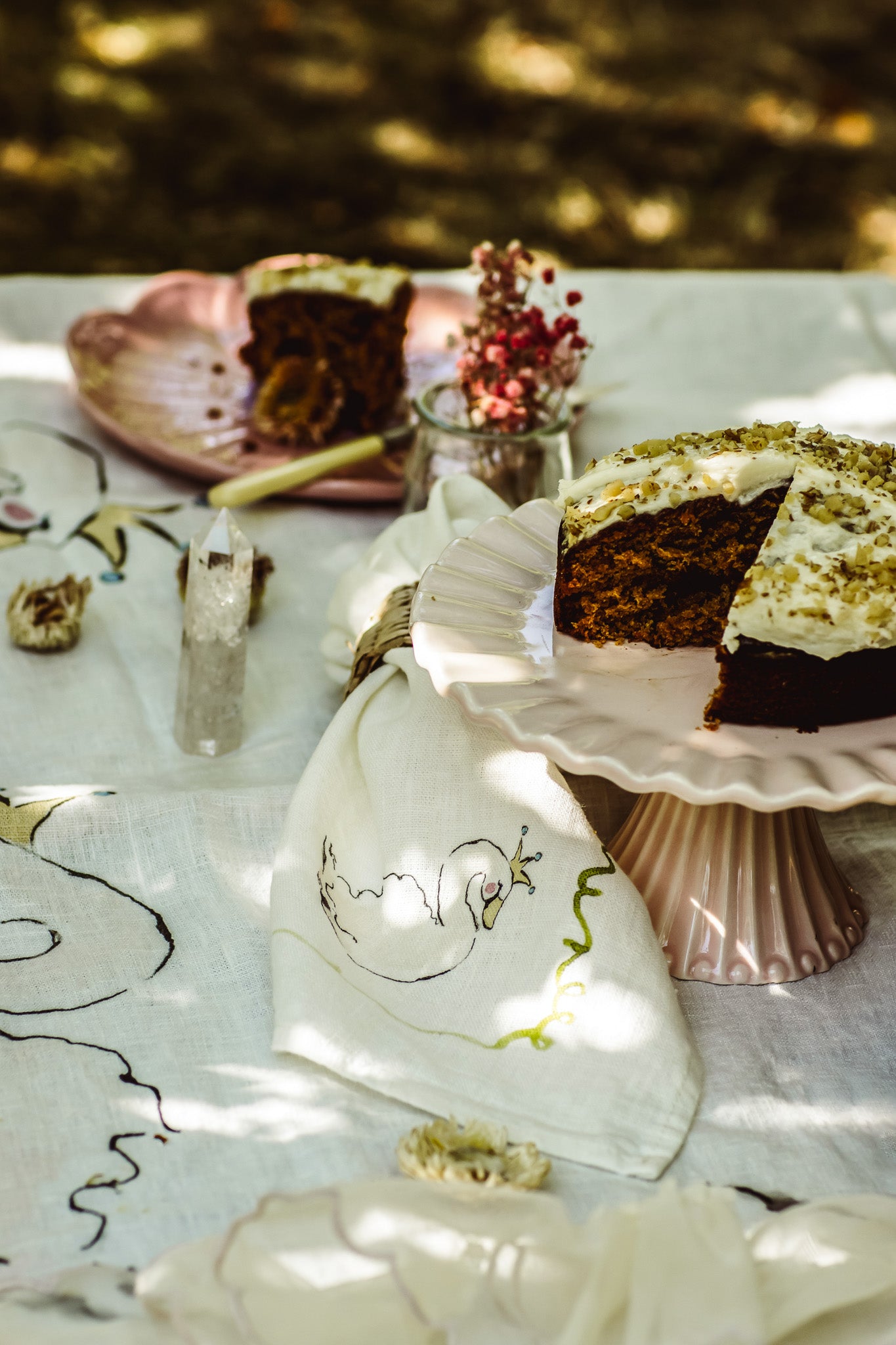 A slice of layered carrot cake with white frosting sits on a pink pedestal plate on a white tablecloth outdoors. Another slice is on a pink plate in the background, with a small vase of dried flowers and a glass candle holder nearby.
