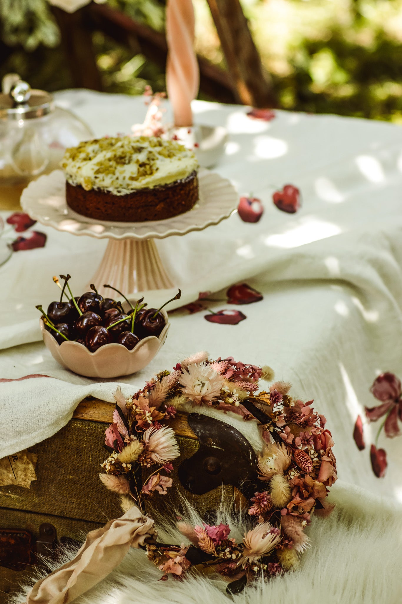 A styled tabletop scene with a decorated cake on a pedestal stand, a bowl of dark cherries, and a dried floral wreath in the foreground on a white cloth outdoors.