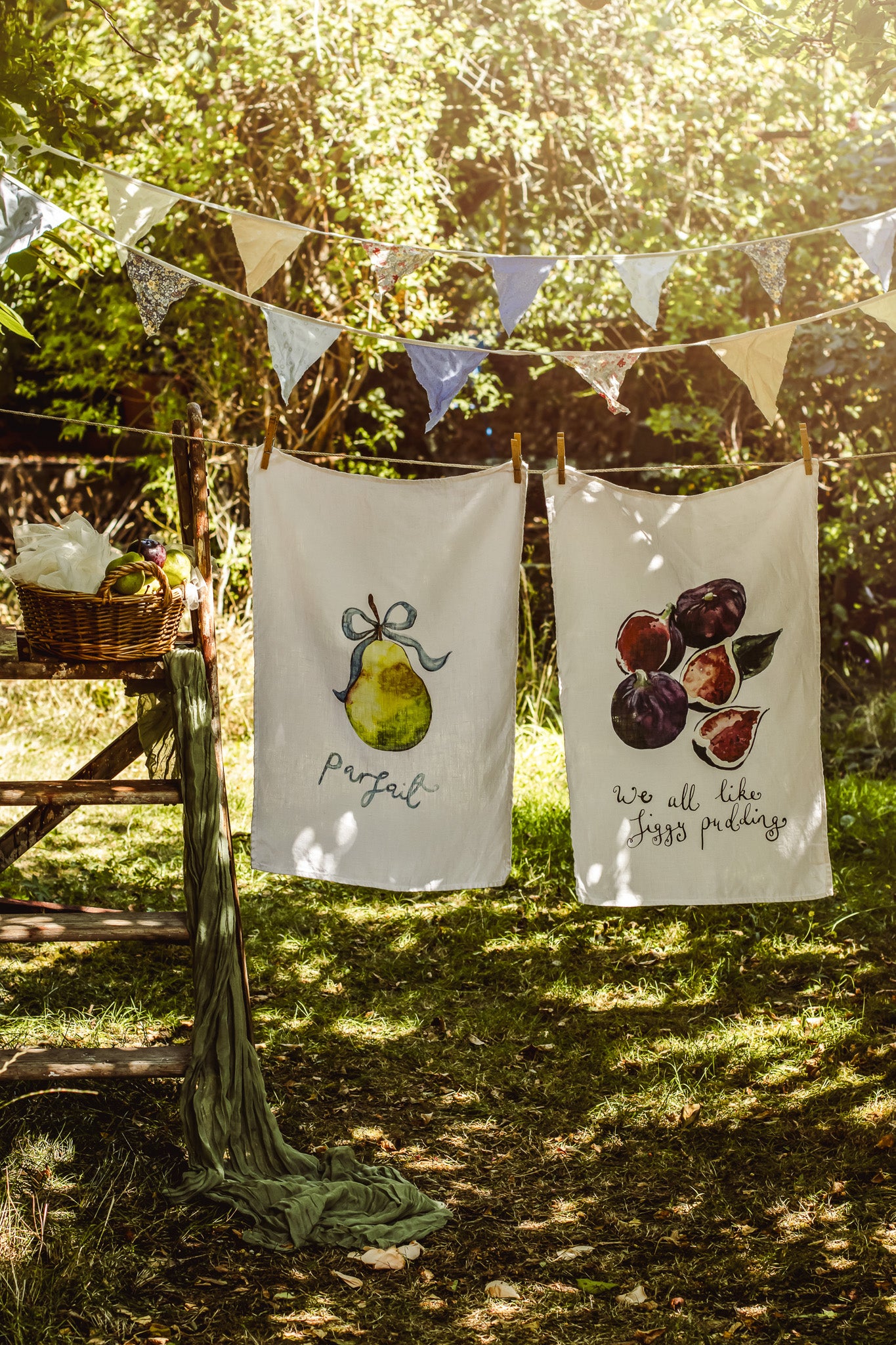 Two white tea towels with watercolor illustrations hanging on a rustic clothesline outdoors in a sunlit garden. The left towel features a green pear with the word 'perfect' underneath, and the right towel shows plums and figs with the phrase 'we all like figs pudding'.