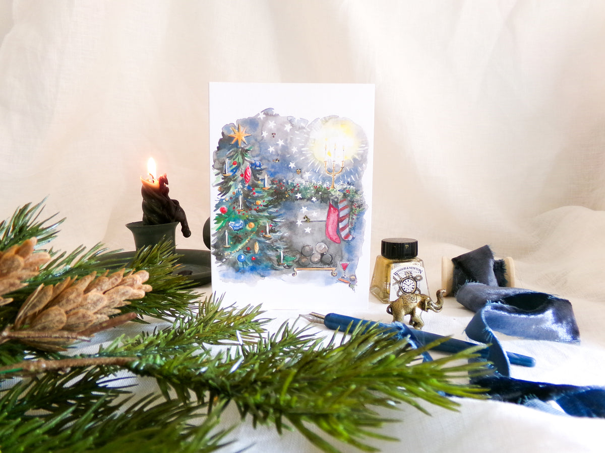 A square art print featuring a whimsical watercolor Christmas scene with a decorated tree, snow, and a glowing light, styled on a white fabric background with pine branches, a candle, and ink as props.