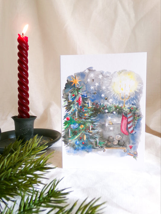 A Christmas-themed art print card featuring a watercolor illustration of a decorated Christmas tree, a glowing candelabra, and a stocking, styled next to a lit red candle in a black holder with green pine branches in the foreground on a white fabric background.