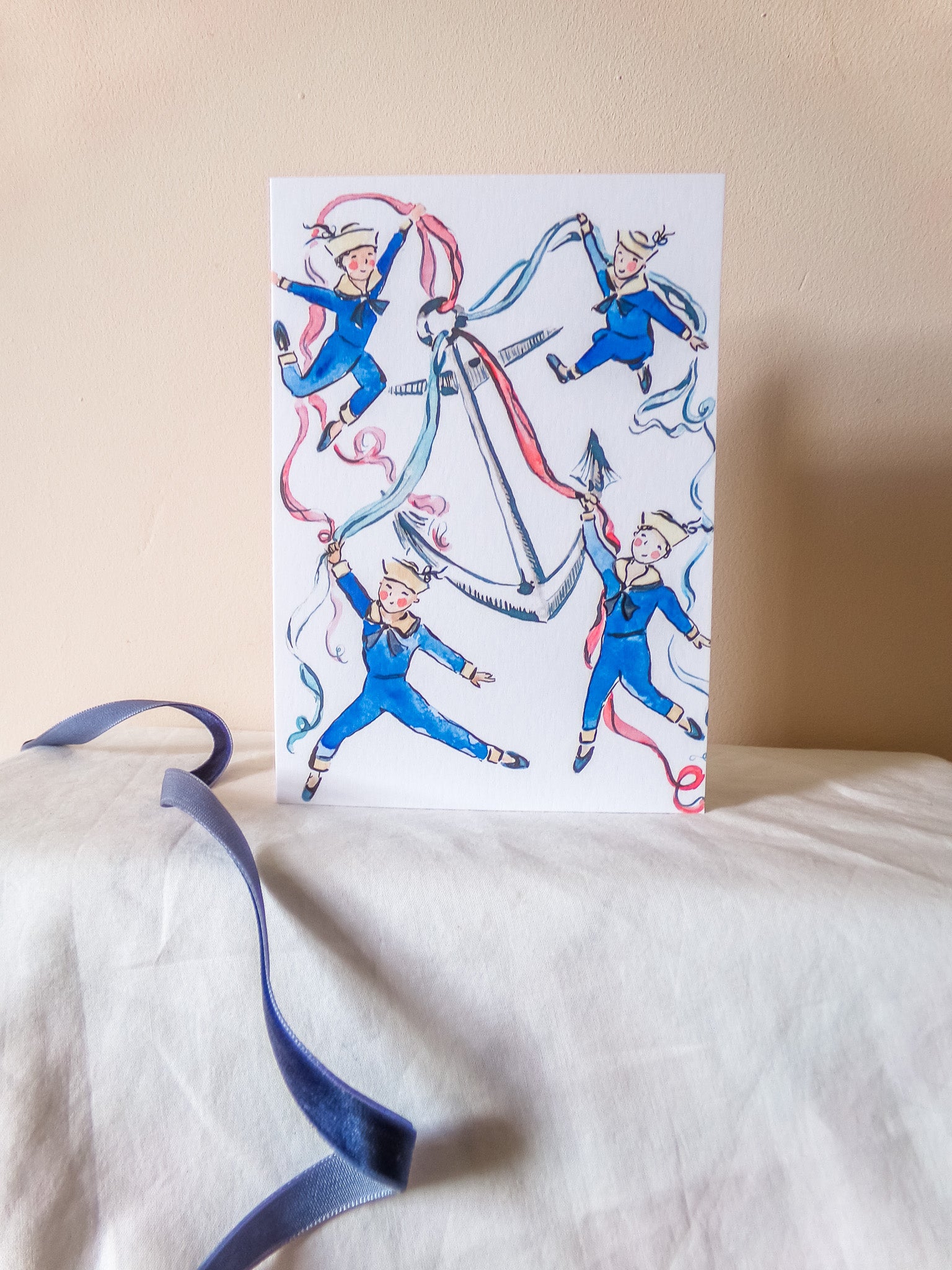 A white greeting card featuring a whimsical illustration of four children in blue uniforms with red, white, and blue ribbons, displayed on a white fabric surface against a beige wall.